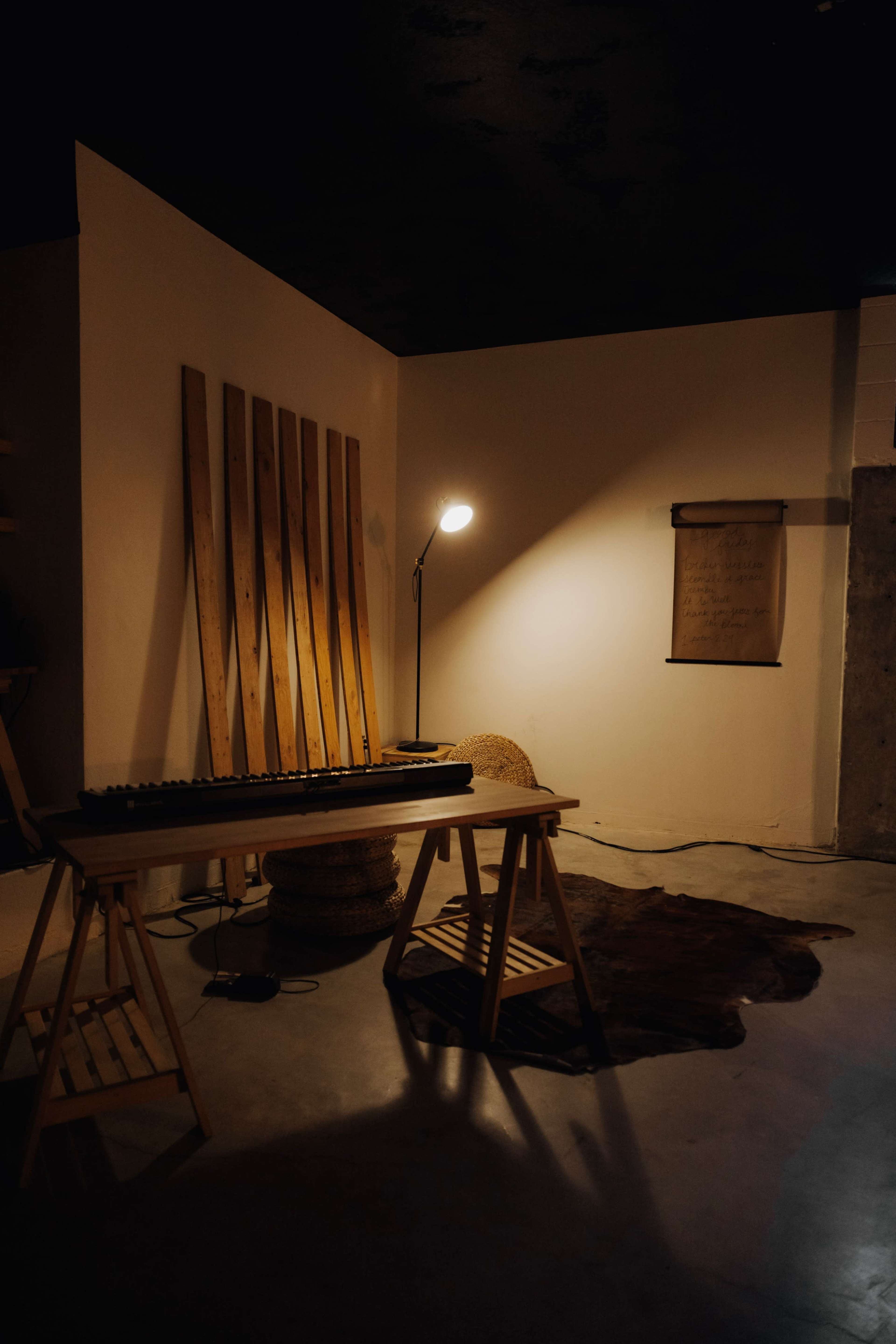 A minimalist room features a wooden table with a keyboard, a floor lamp, and a scroll hanging on the wall, accompanied by woven stools and a rug on the concrete floor.