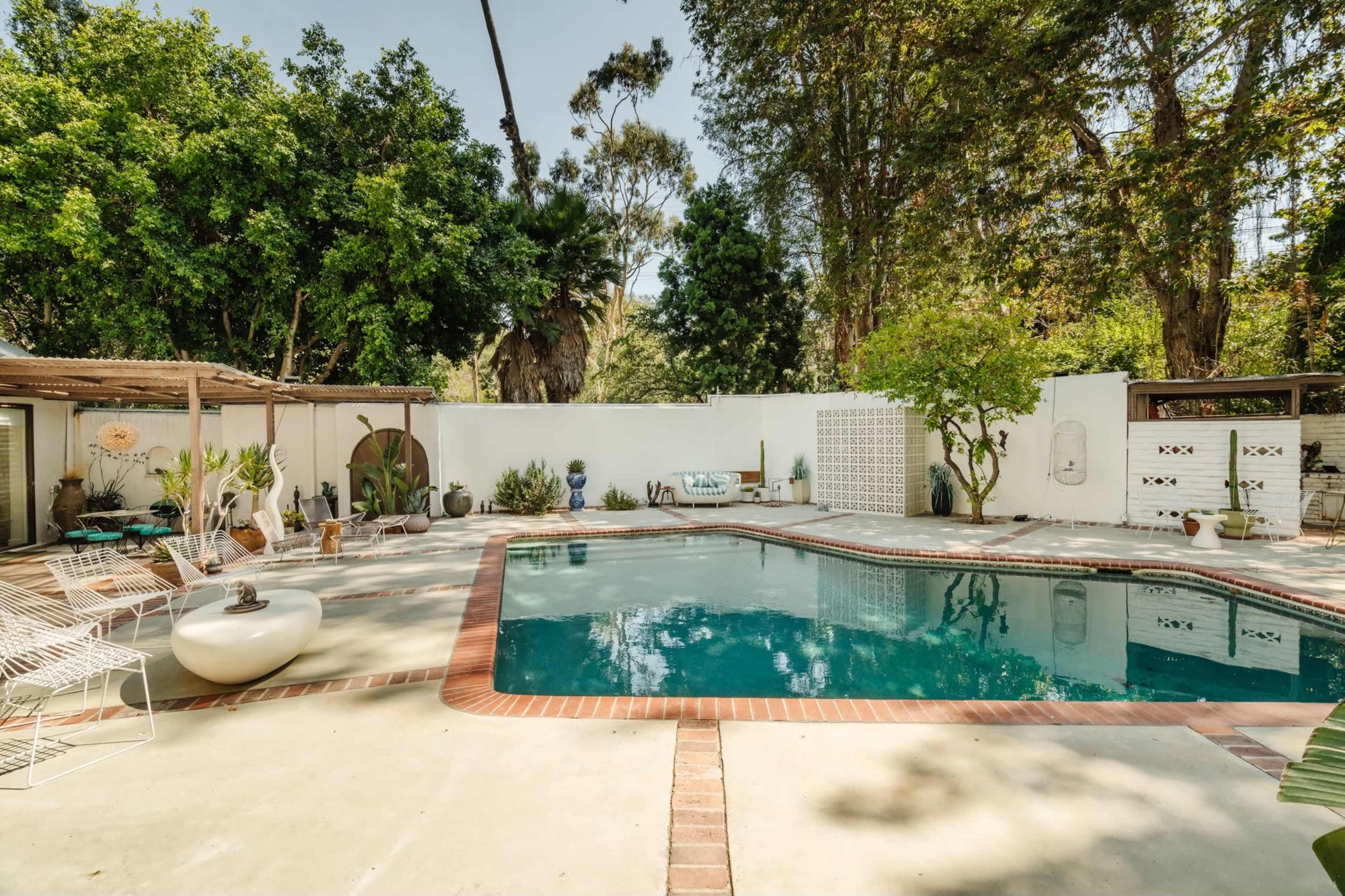 The image shows a tranquil outdoor space featuring a rectangular swimming pool surrounded by a paved deck, plants, and outdoor furniture.