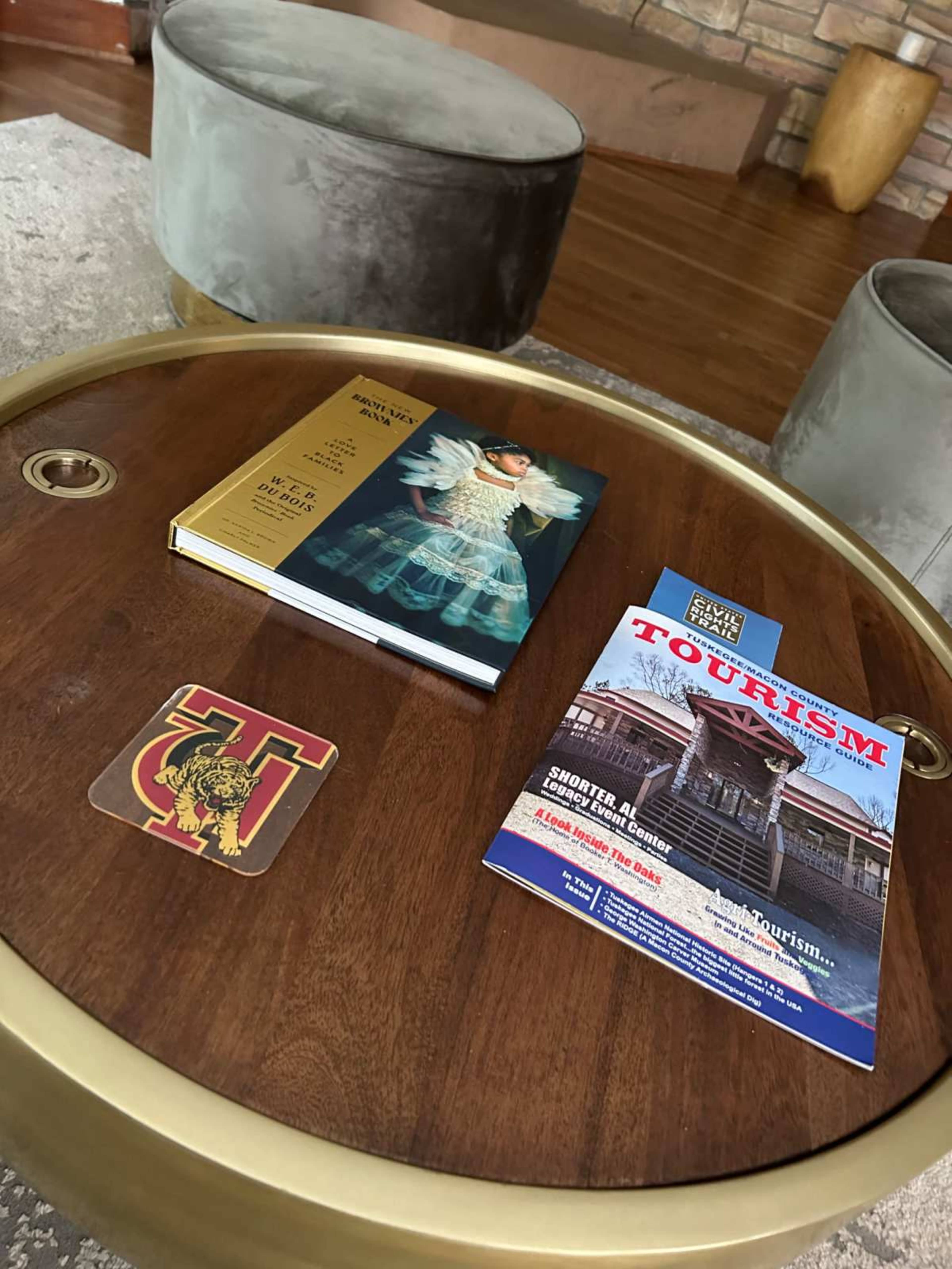A round wooden coffee table displays a magazine, a brochure, and a book, along with a coaster featuring a logo.