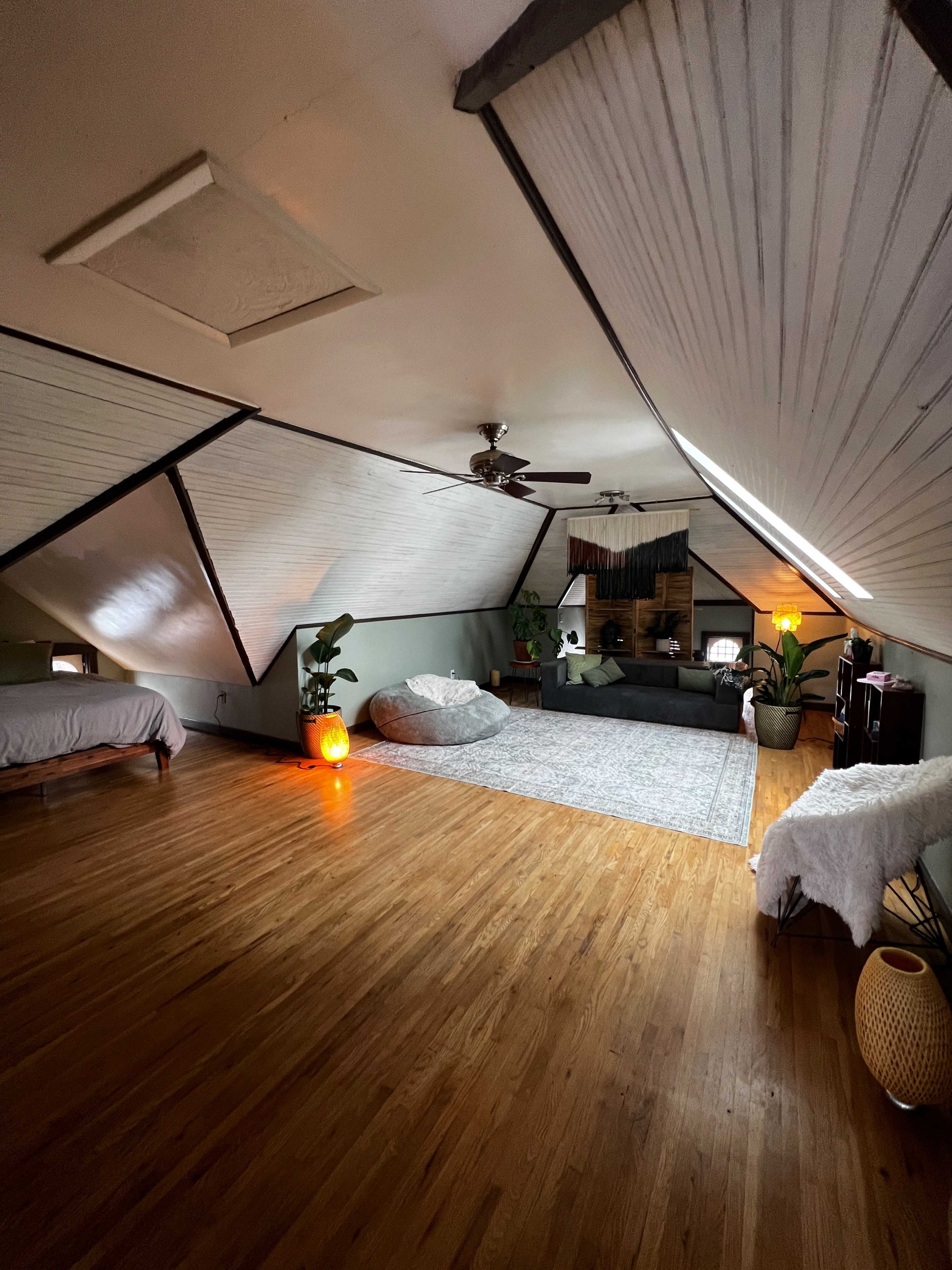 The image shows a spacious loft-style room with wooden floors, a large area rug, and various seating options, including a bean bag and two beds, under slanted ceilings.