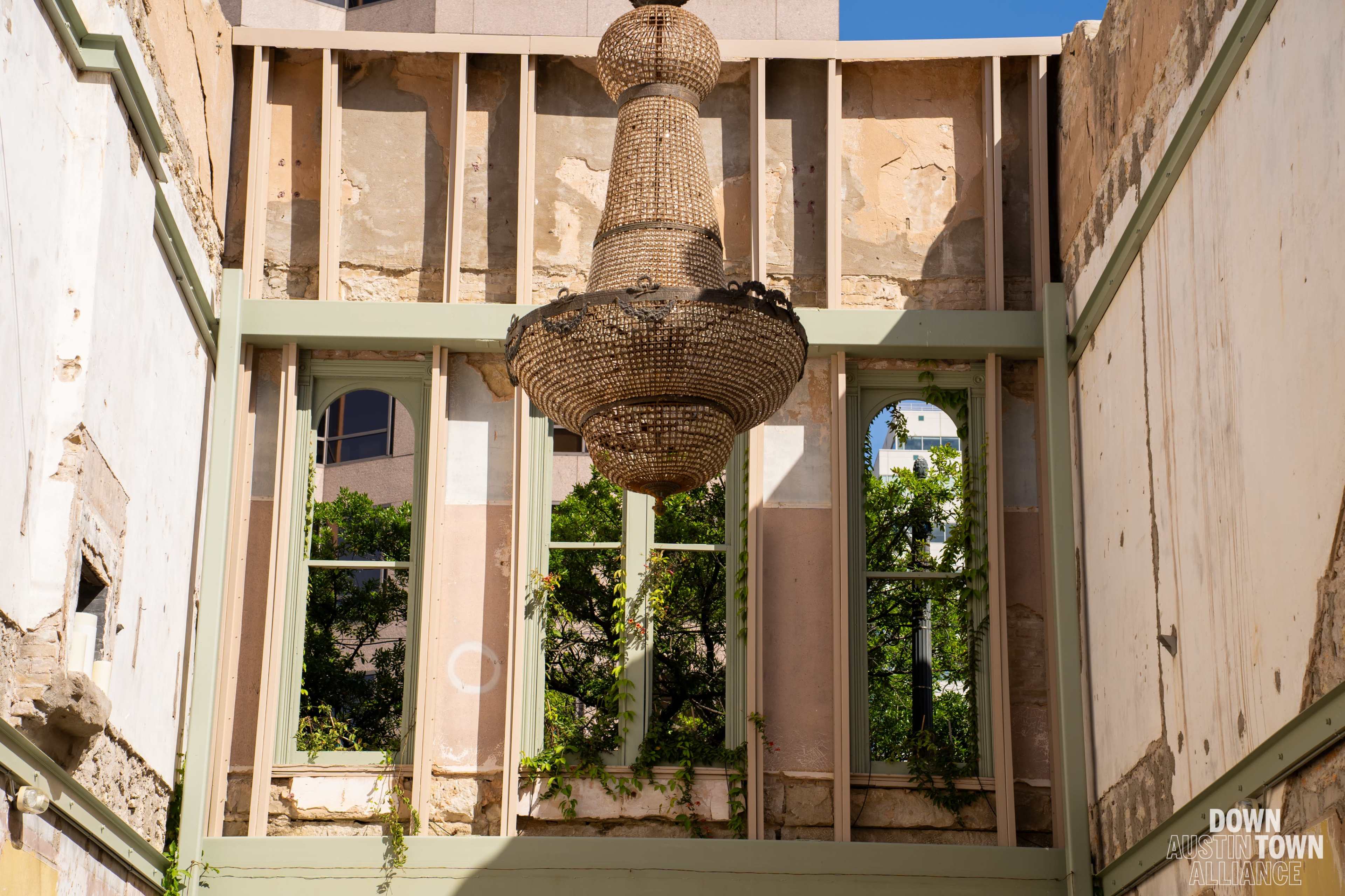 A large, woven chandelier hangs from the ceiling of a dilapidated interior, while shattered walls reveal green foliage outside.