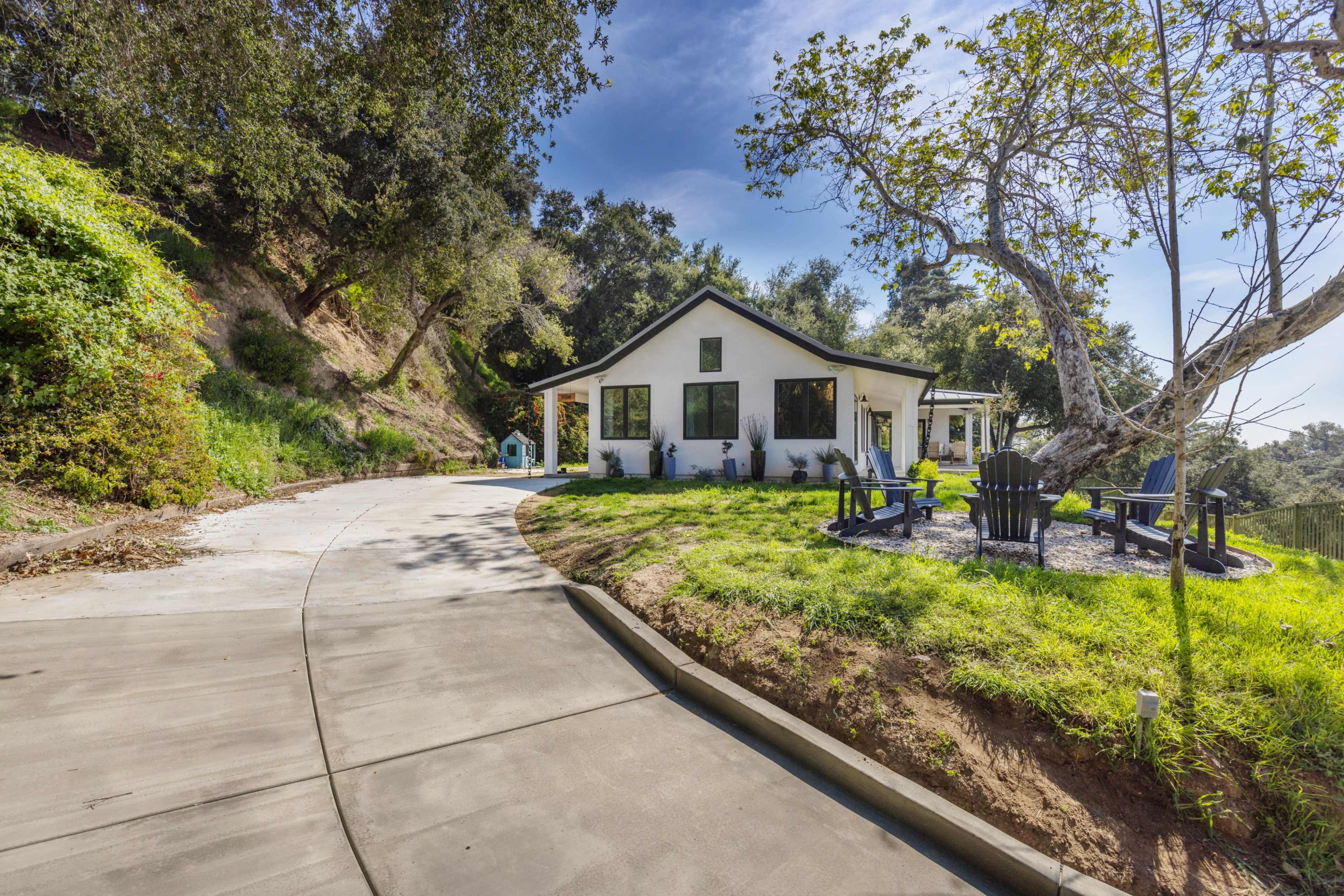 A modern house sits on a hillside, surrounded by trees, with a curved driveway leading to the front yard featuring outdoor seating.