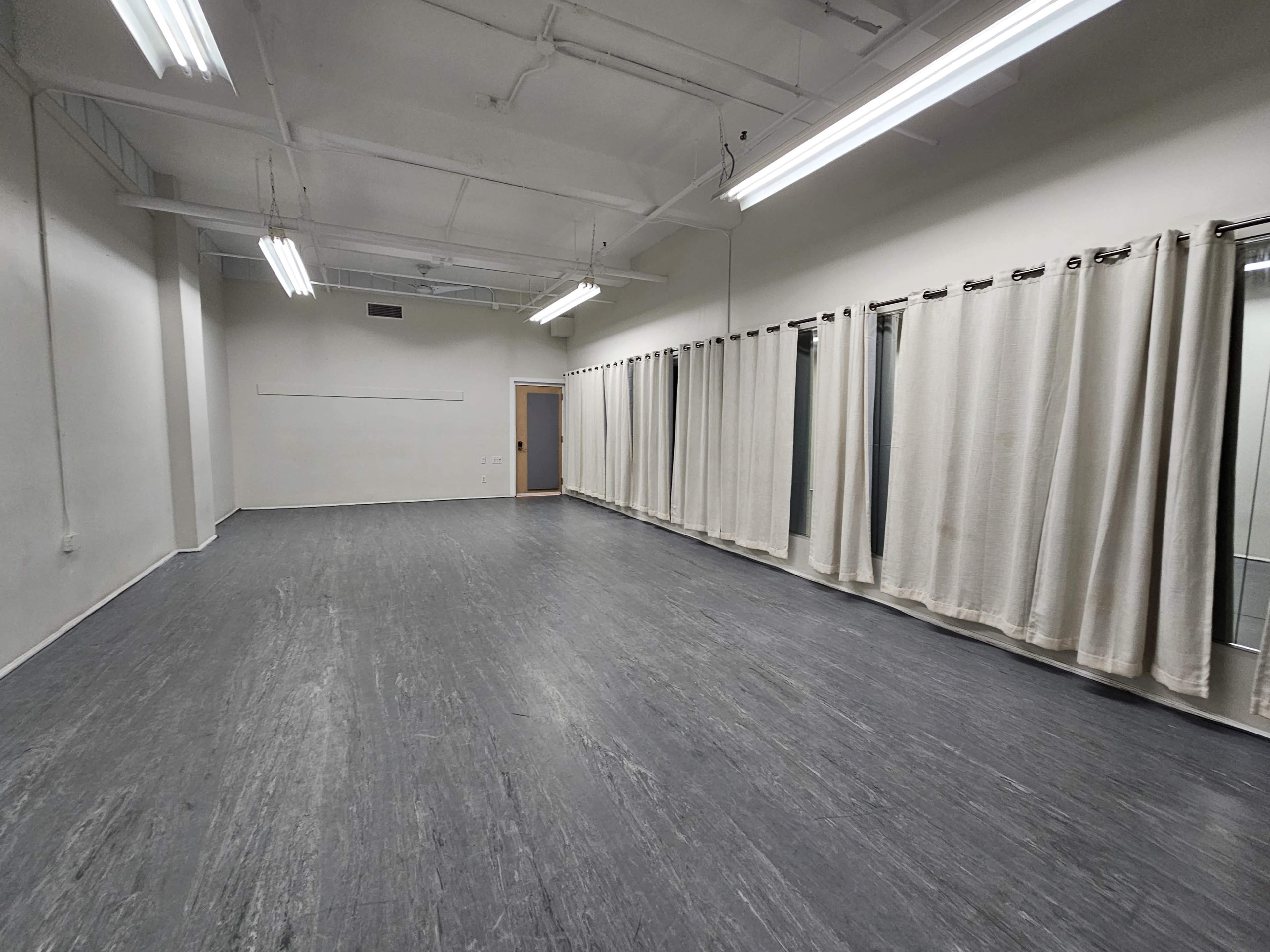 A spacious, empty room with gray flooring, white walls, and large windows covered by curtains.