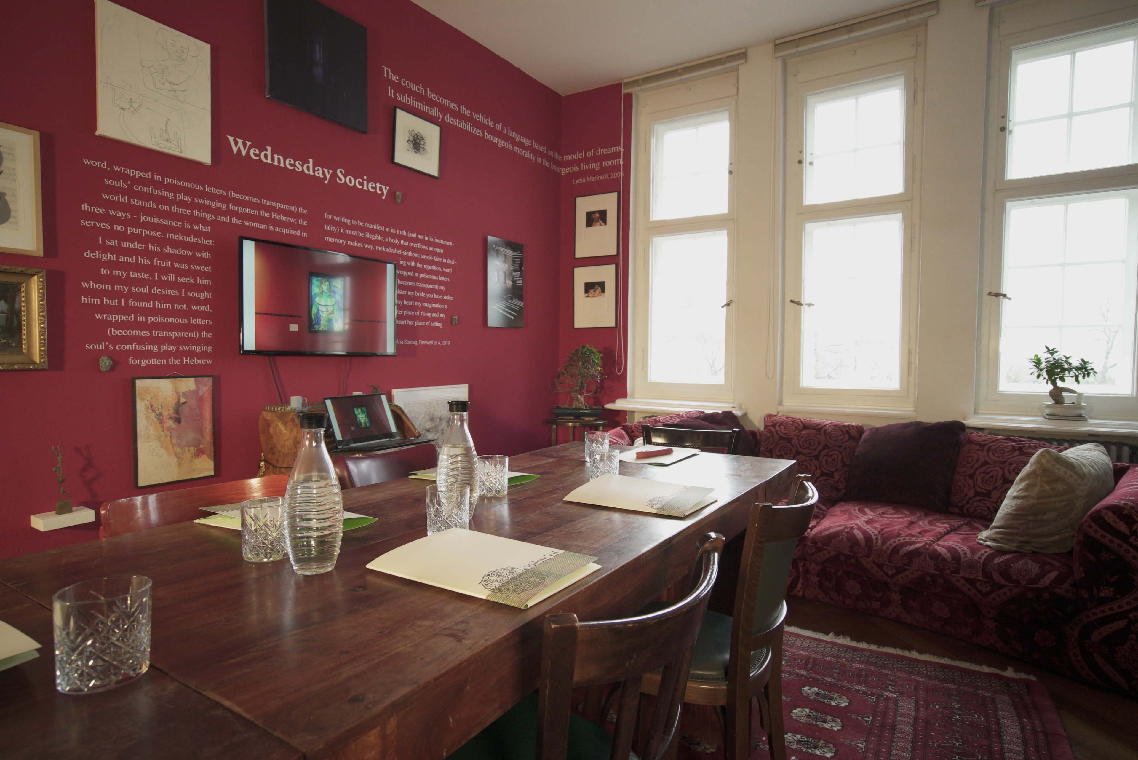 A dining room features a long wooden table set with glassware and place settings, against a red wall adorned with framed artwork and text.