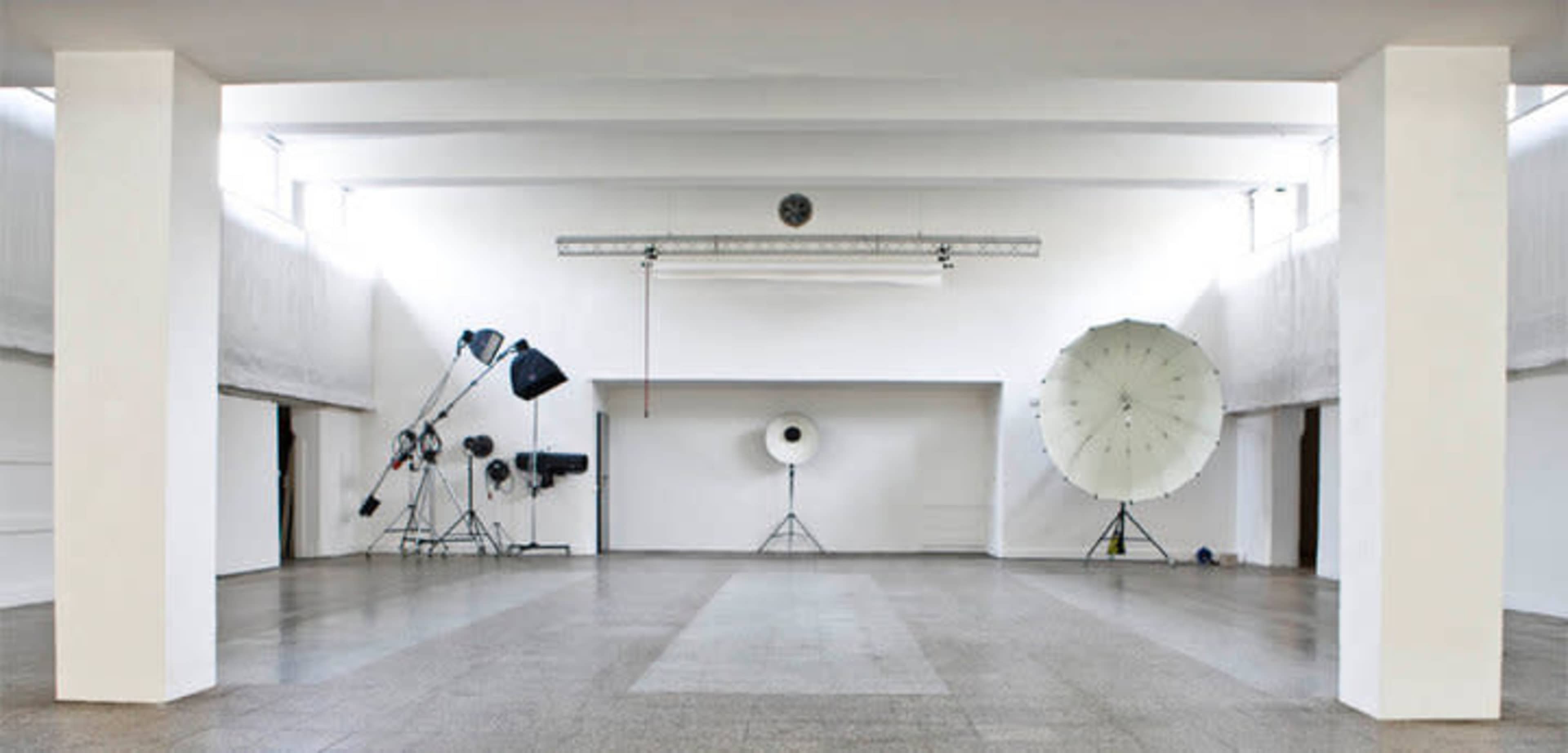 A large, empty studio space with white walls and various photography equipment, including light stands and large reflectors, arranged in the corners.