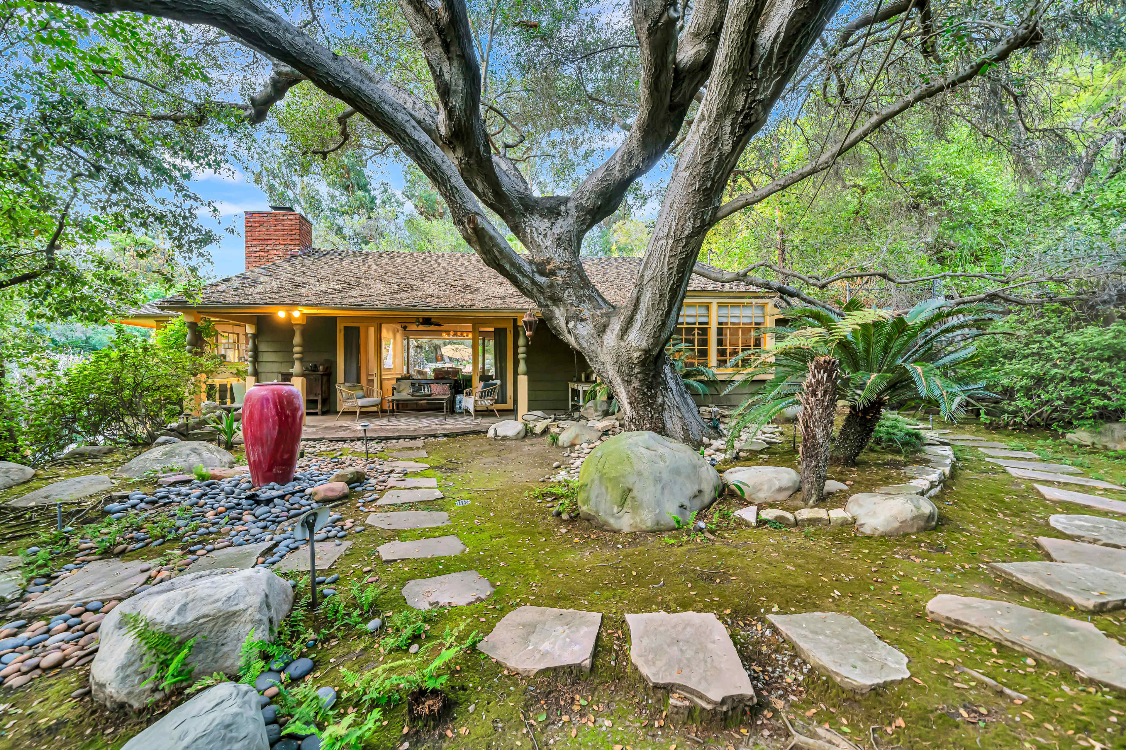 A cozy wooden cottage is surrounded by lush greenery, featuring a stone pathway and a large tree in the yard.