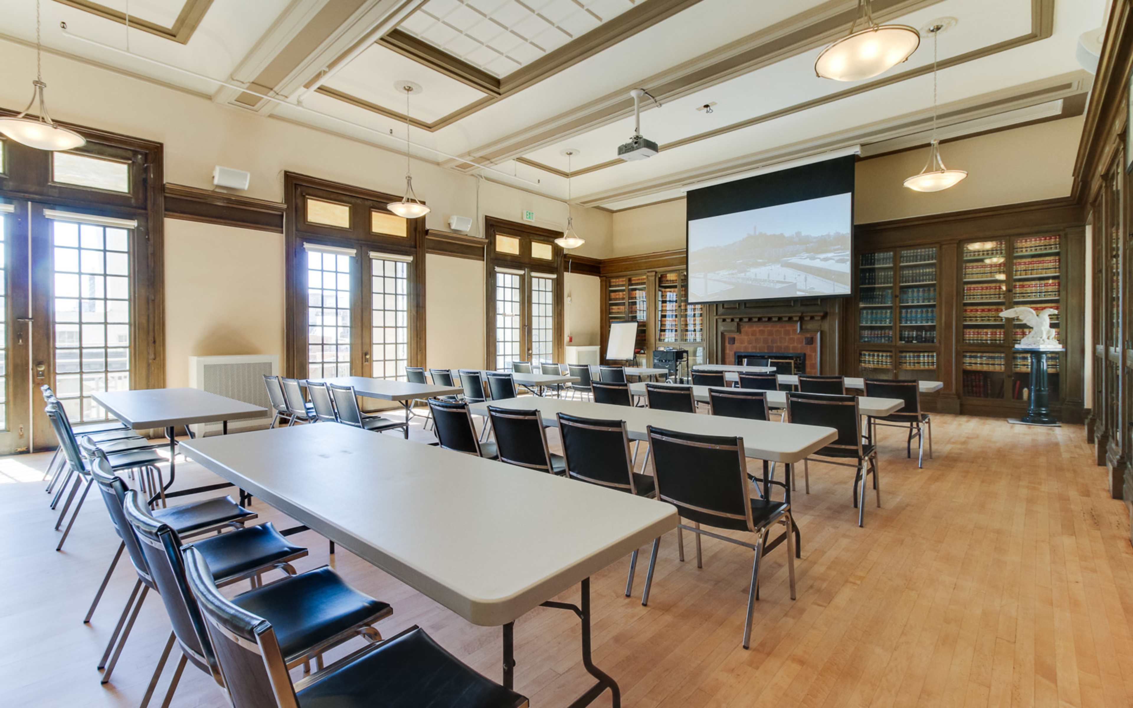 A seminar room is set up with rows of tables and chairs facing a large screen, featuring large windows and bookshelves along the walls.