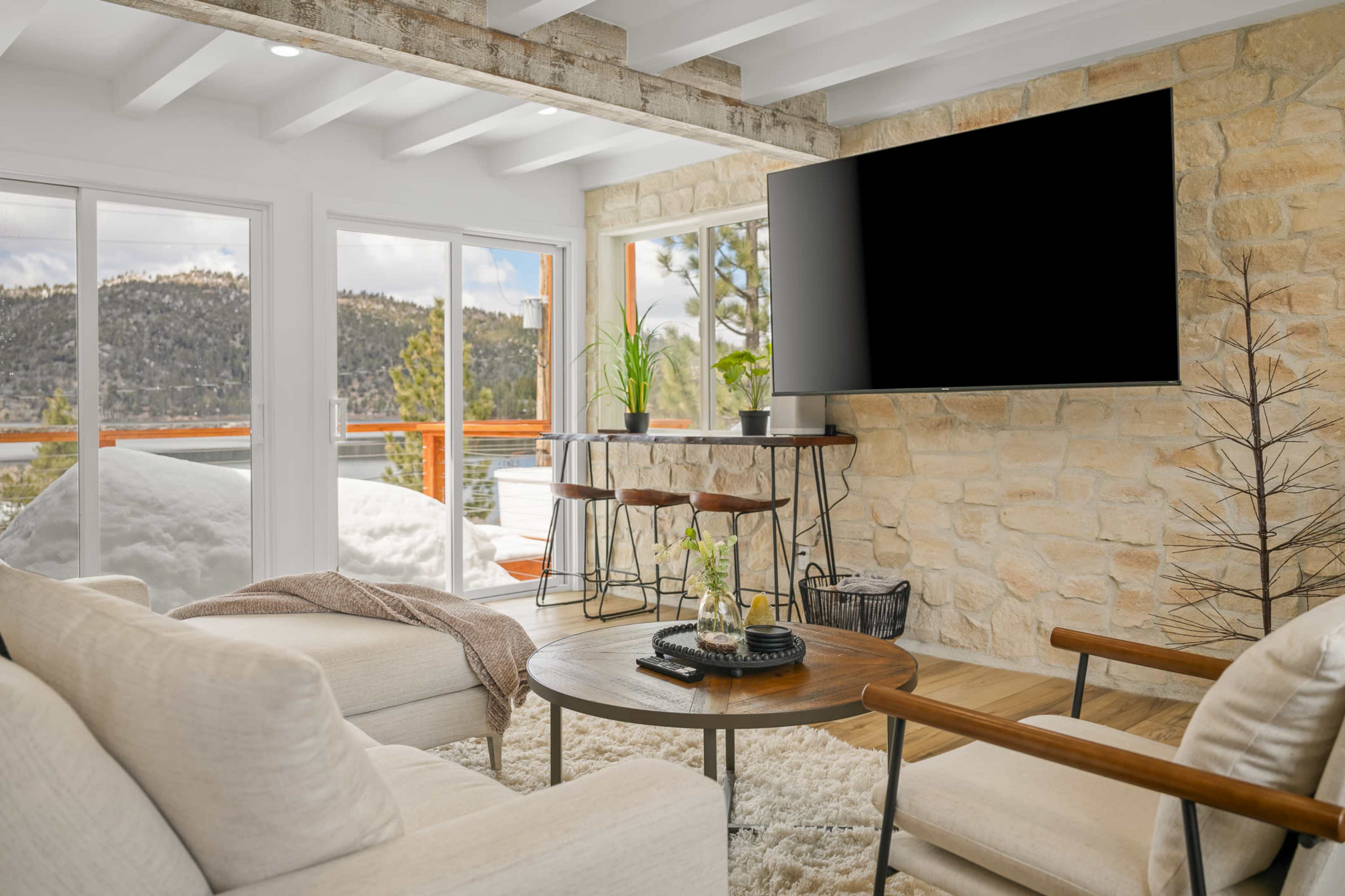 The living room features a stone wall, a large television mounted above a circular table, and seating arranged to face expansive windows overlooking a snowy landscape.