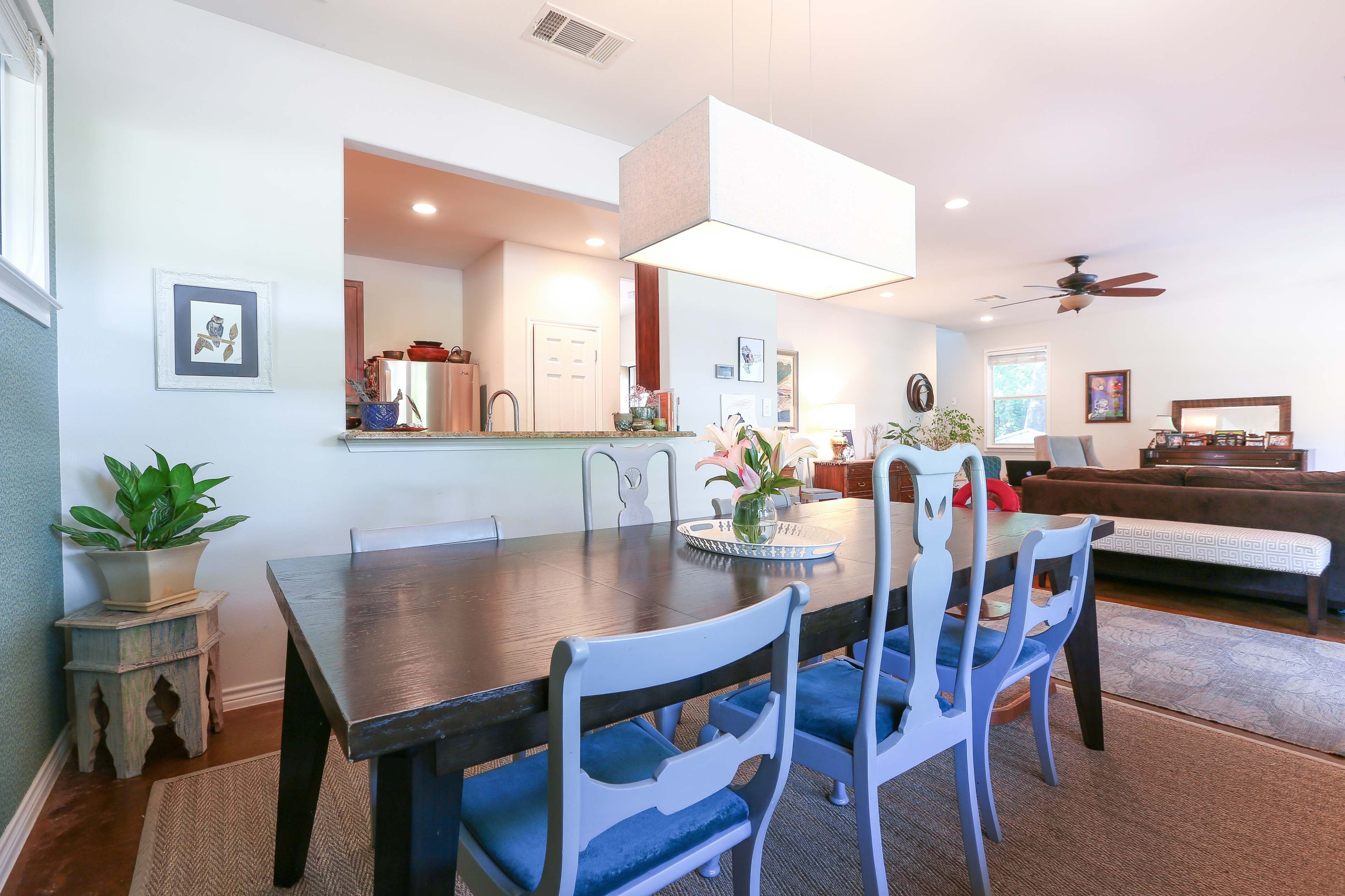 A dining area features a wooden table with six chairs, a large light fixture overhead, and an open view into a kitchen and living room space.