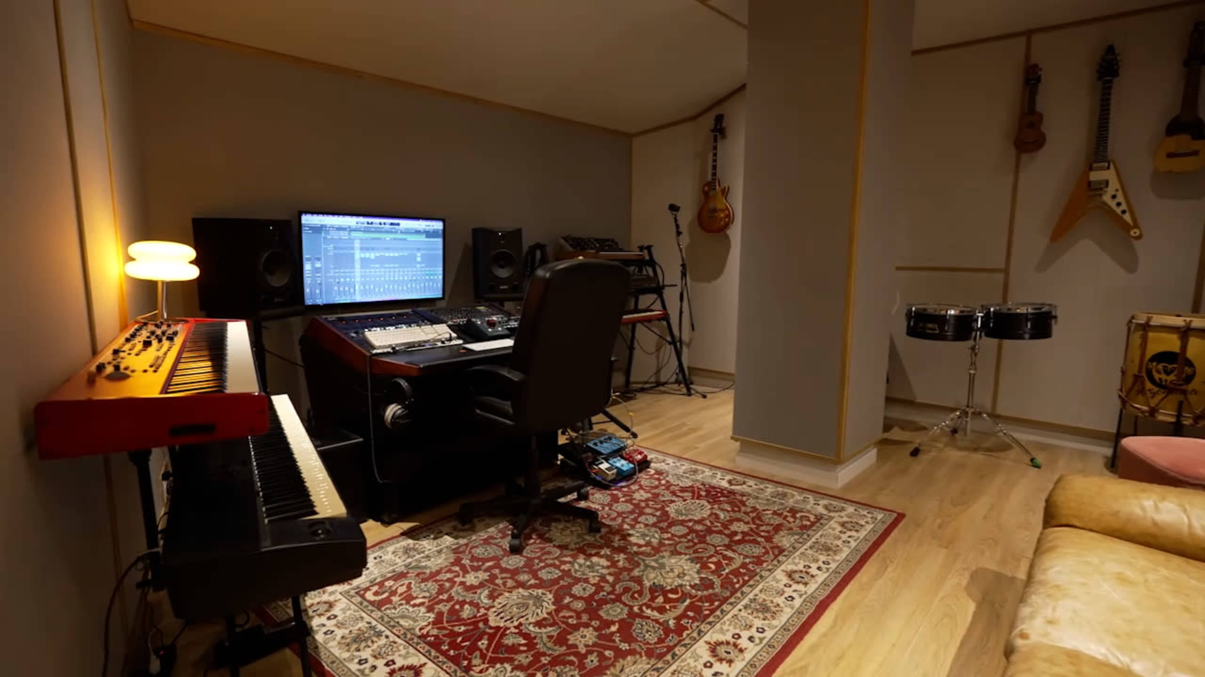 The image shows a home music studio featuring a computer setup, keyboard, and various musical instruments on the walls.