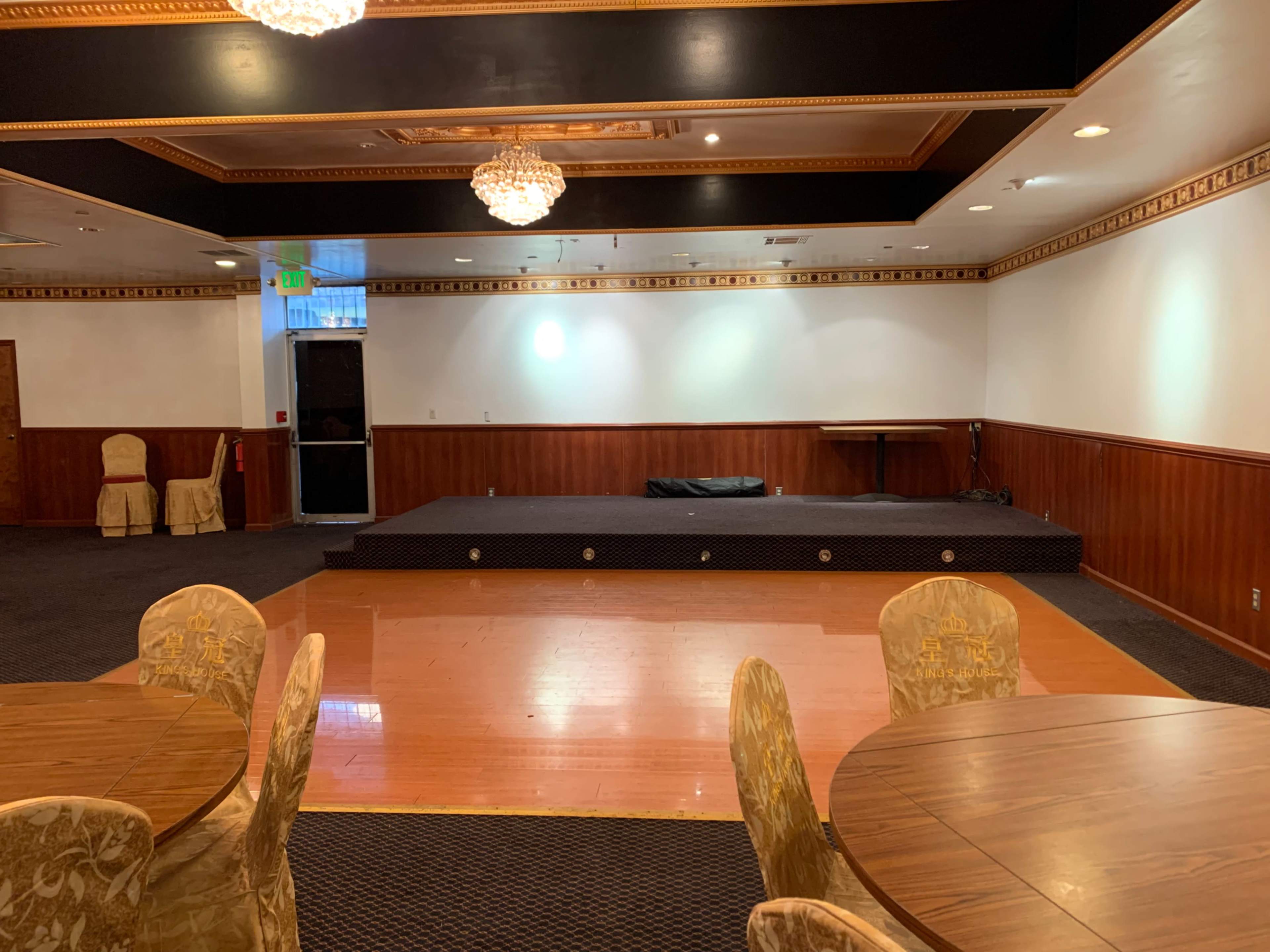 An empty room featuring a raised stage, several round tables, and a wooden floor with a partial view of a partitioned area.