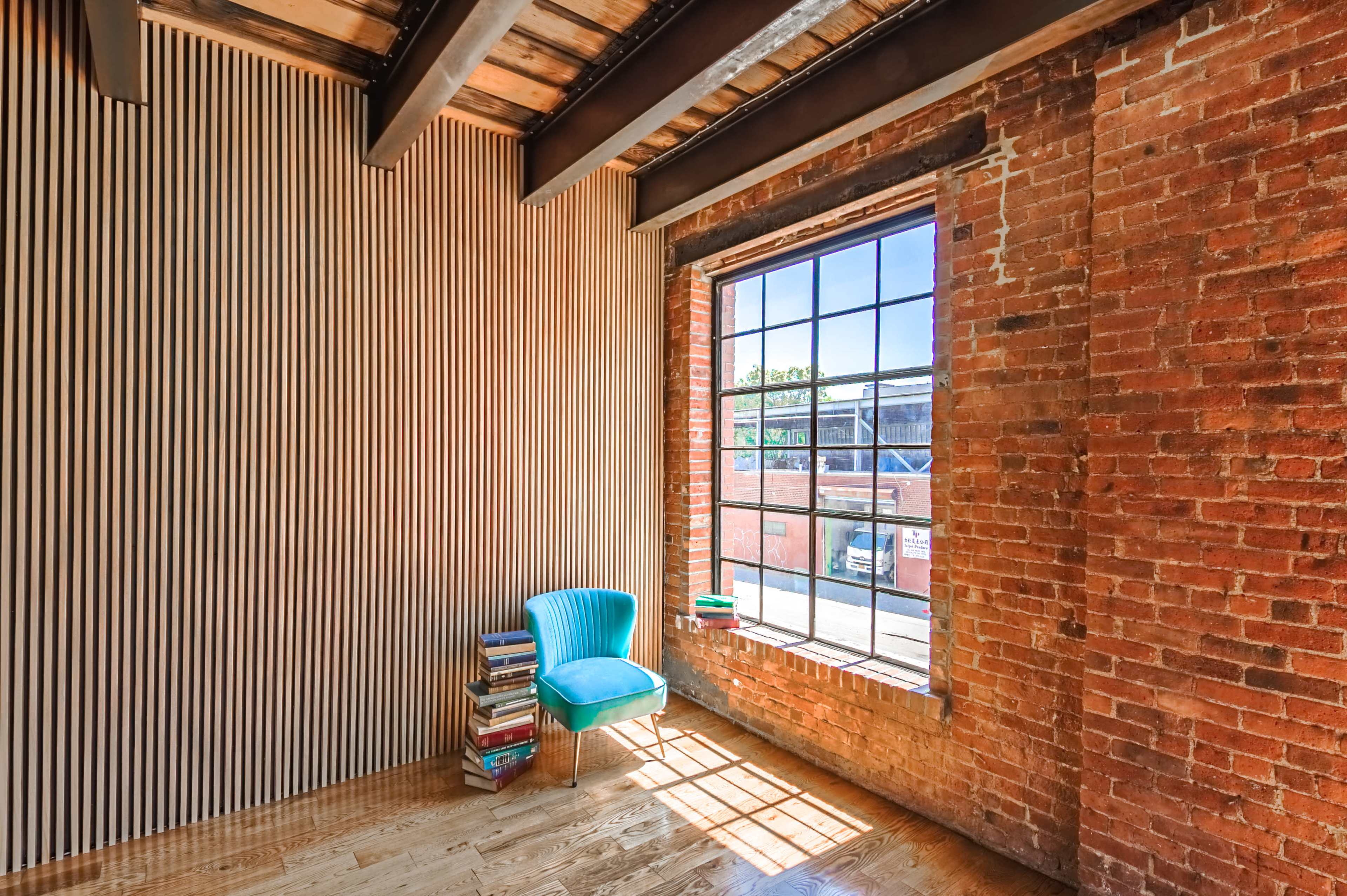 The image shows a bright room with a large window, a turquoise chair, and a stack of books against a brick wall with vertical wooden paneling.