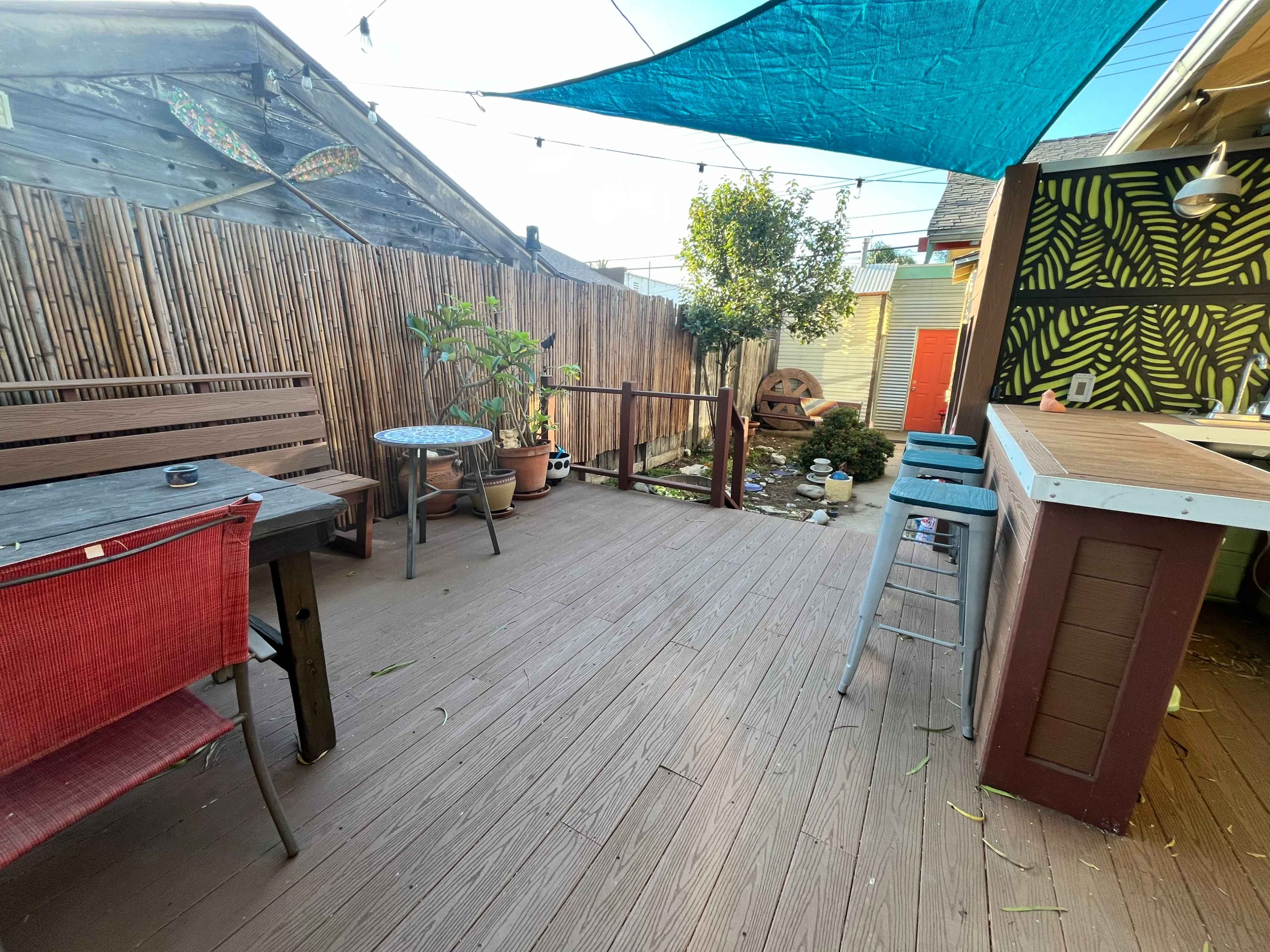The image shows a small outdoor deck area featuring wooden flooring, potted plants, a table with chairs, and a bar counter, all under a blue canopy.