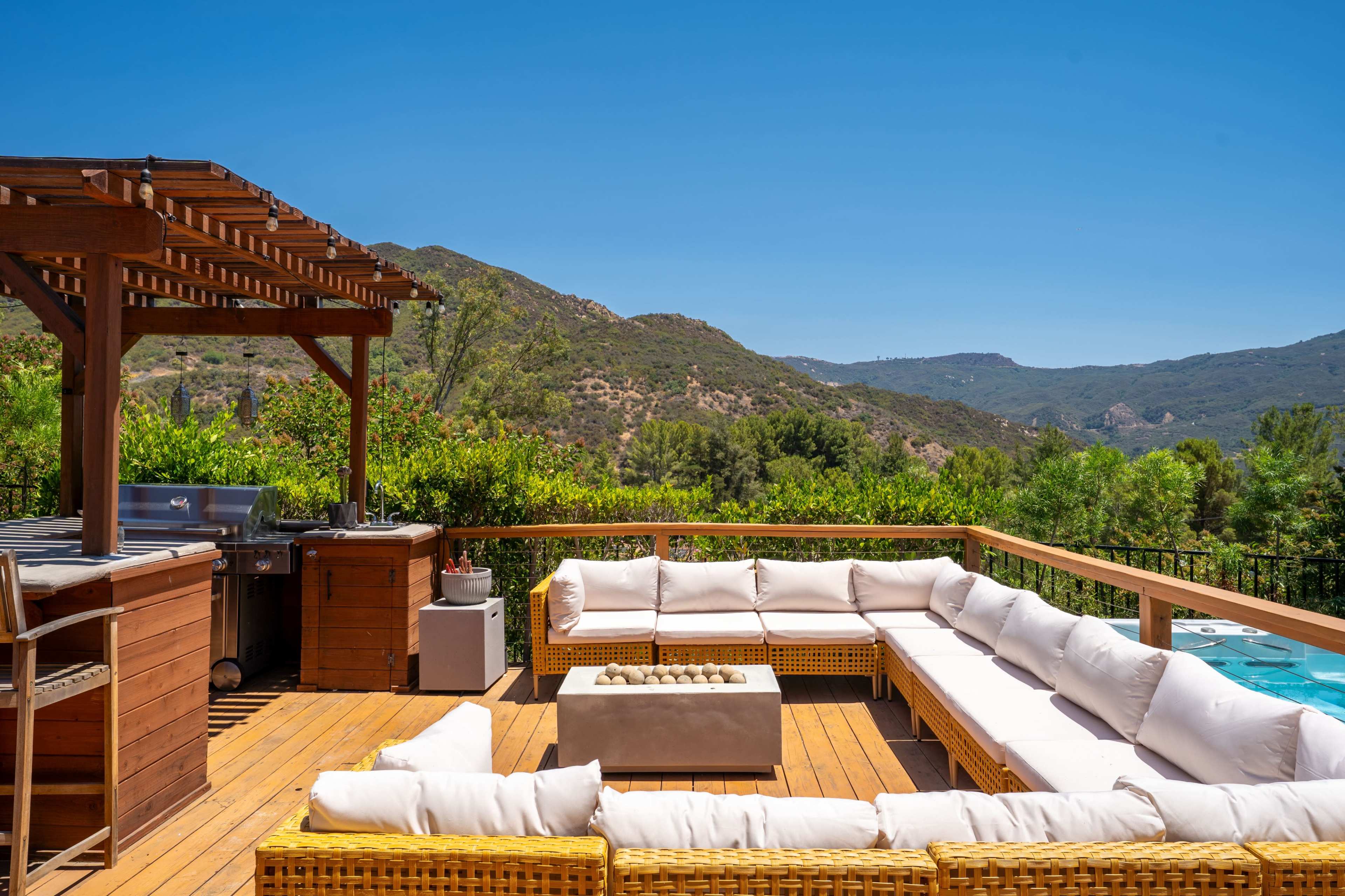 The image features a wooden deck with a large outdoor seating area, a grill, and a hot tub, all set against a backdrop of green hills and clear blue skies.