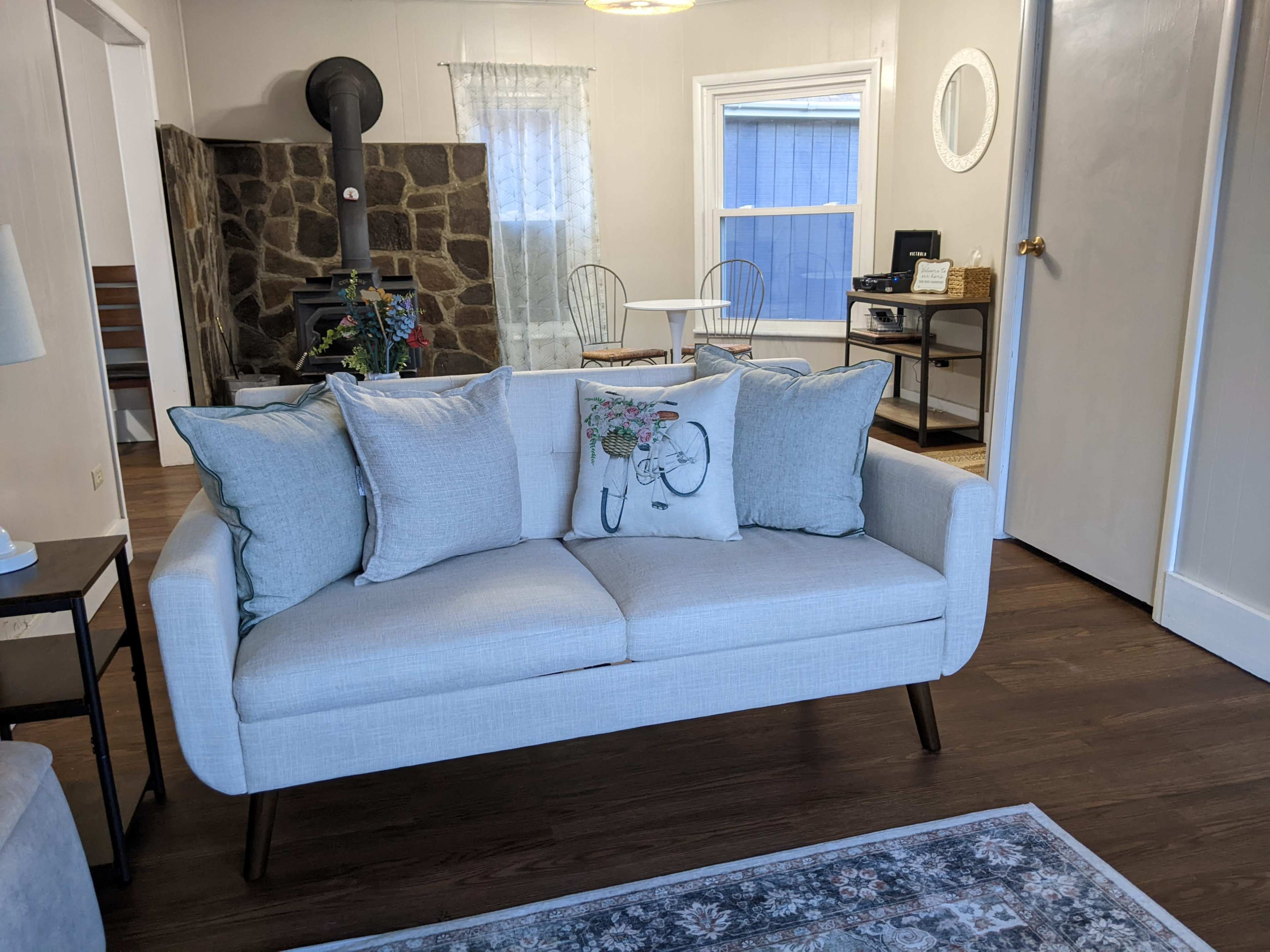 A light-colored sofa with decorative pillows is positioned in a living area that features a stone fireplace and a dining table in the background.