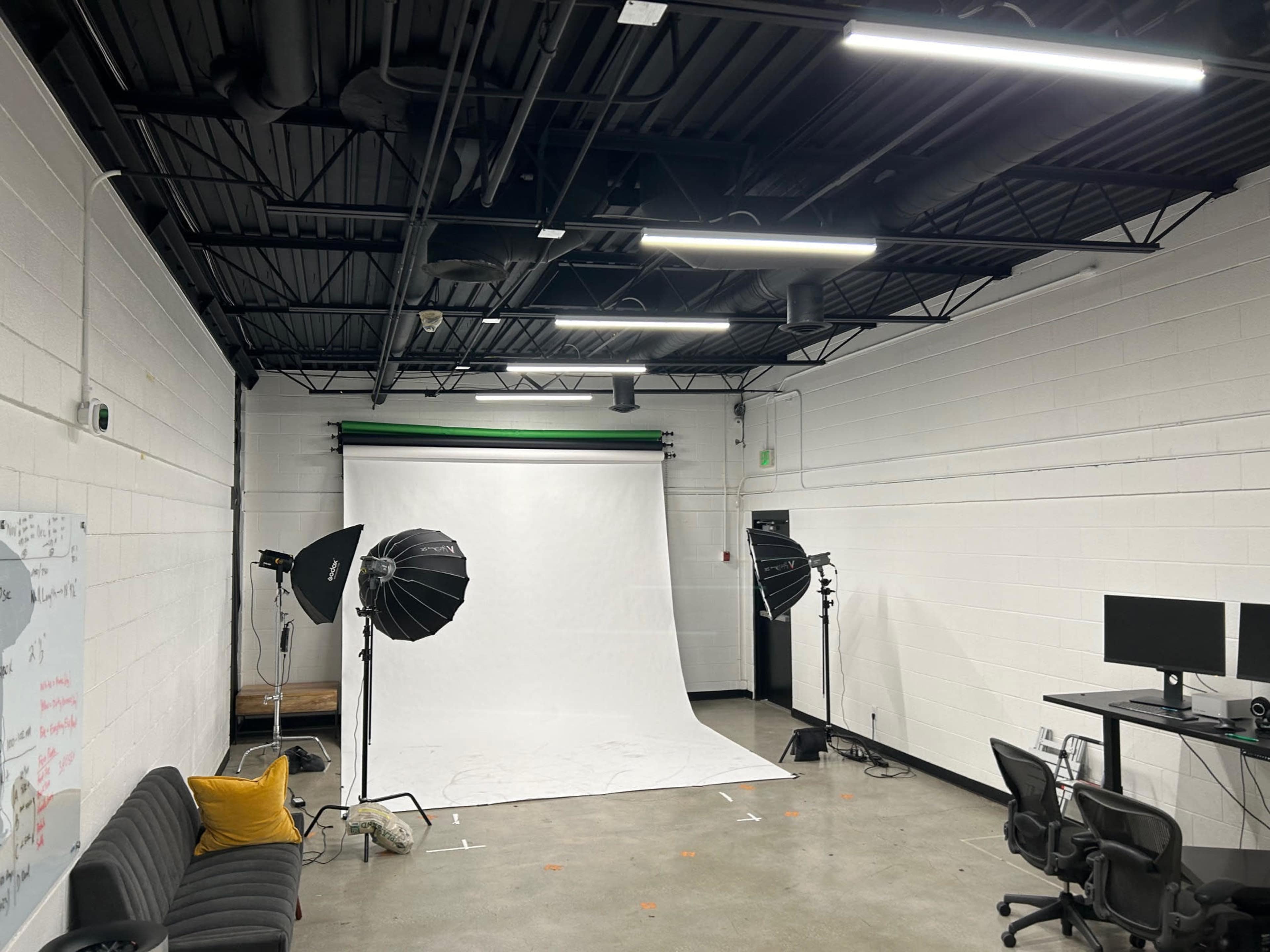 A photography studio with a white backdrop, lighting equipment, a black sofa, and two computer monitors on a desk.