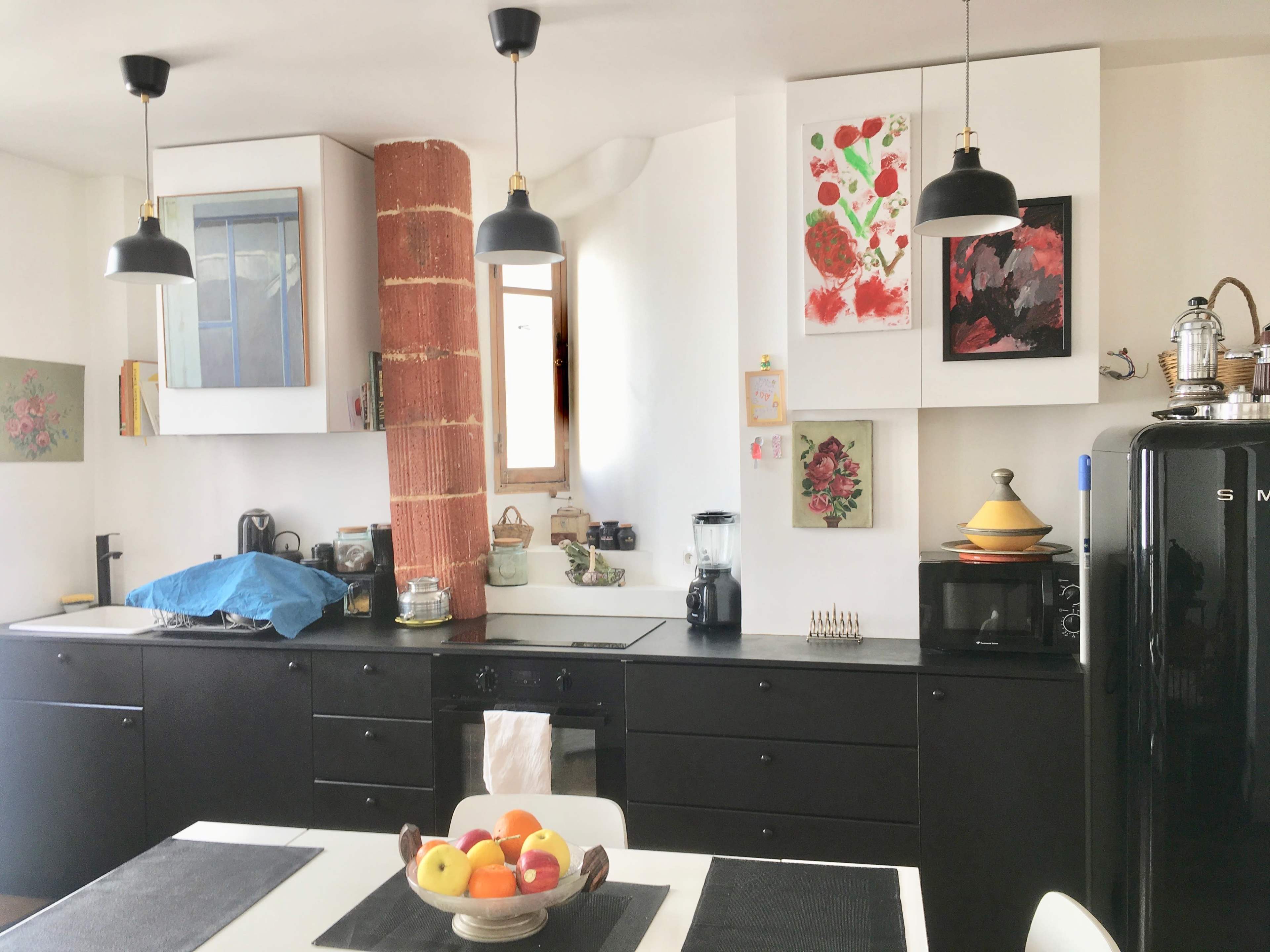 A modern kitchen features black cabinetry, white walls, and a central table with a fruit bowl, illuminated by pendant lamps.