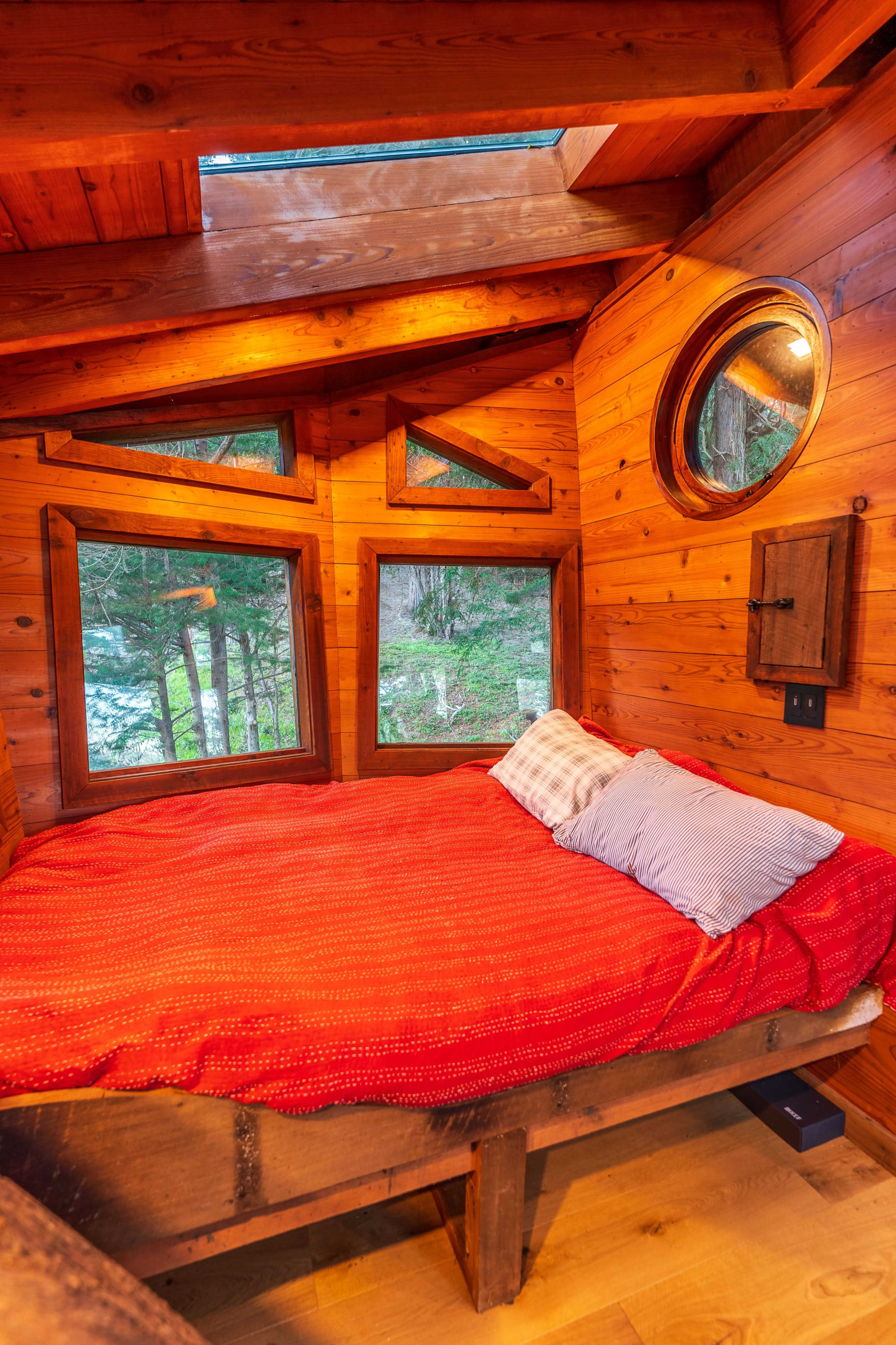 The interior of a wooden cabin features a circular window and a bed with a red blanket, surrounded by wooden walls and multiple angular windows.