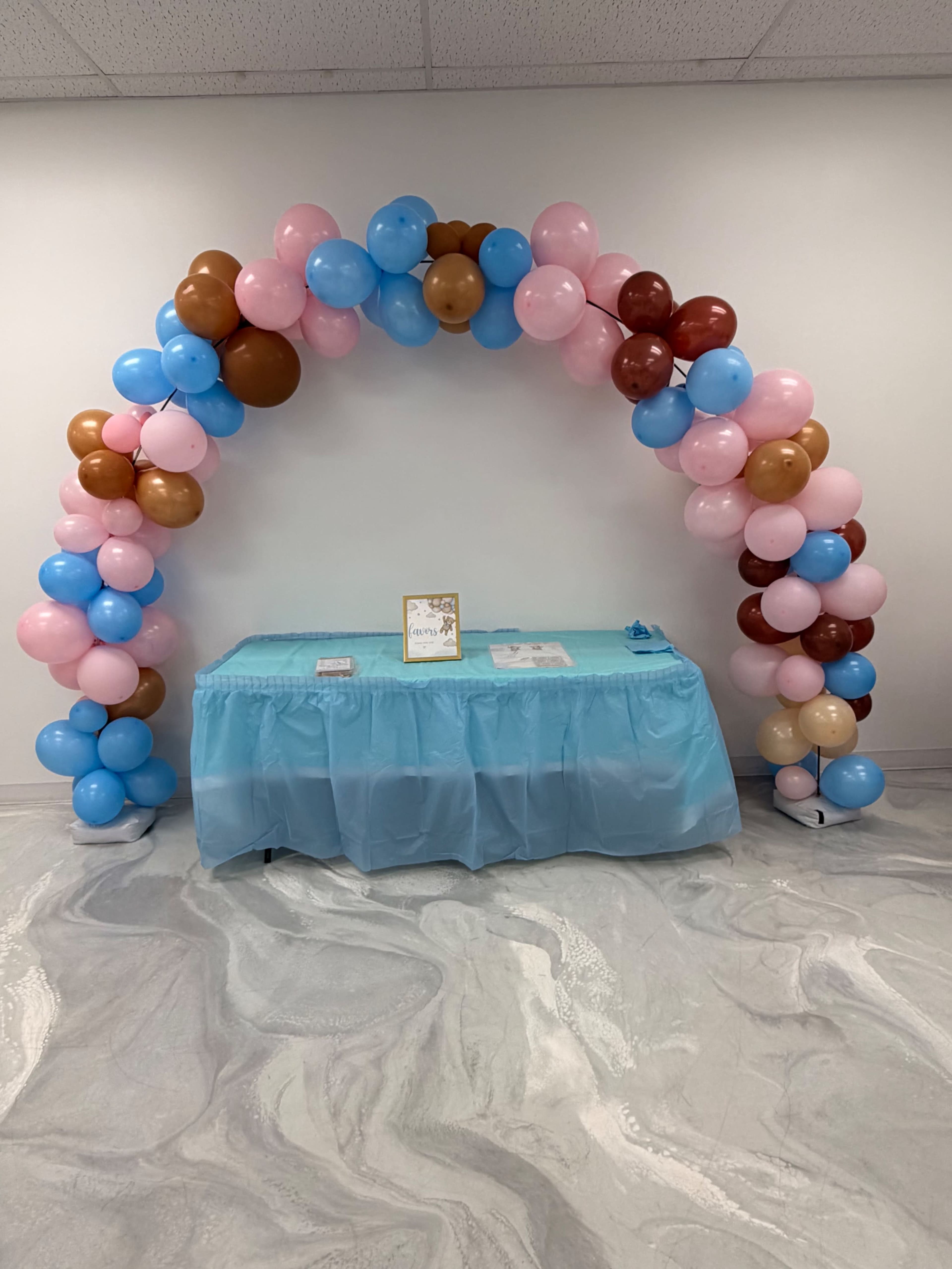 A colorful balloon arch made of pastel blue, pink, brown, and tan balloons stands over a light blue tablecloth set with a decorative sign and additional items.