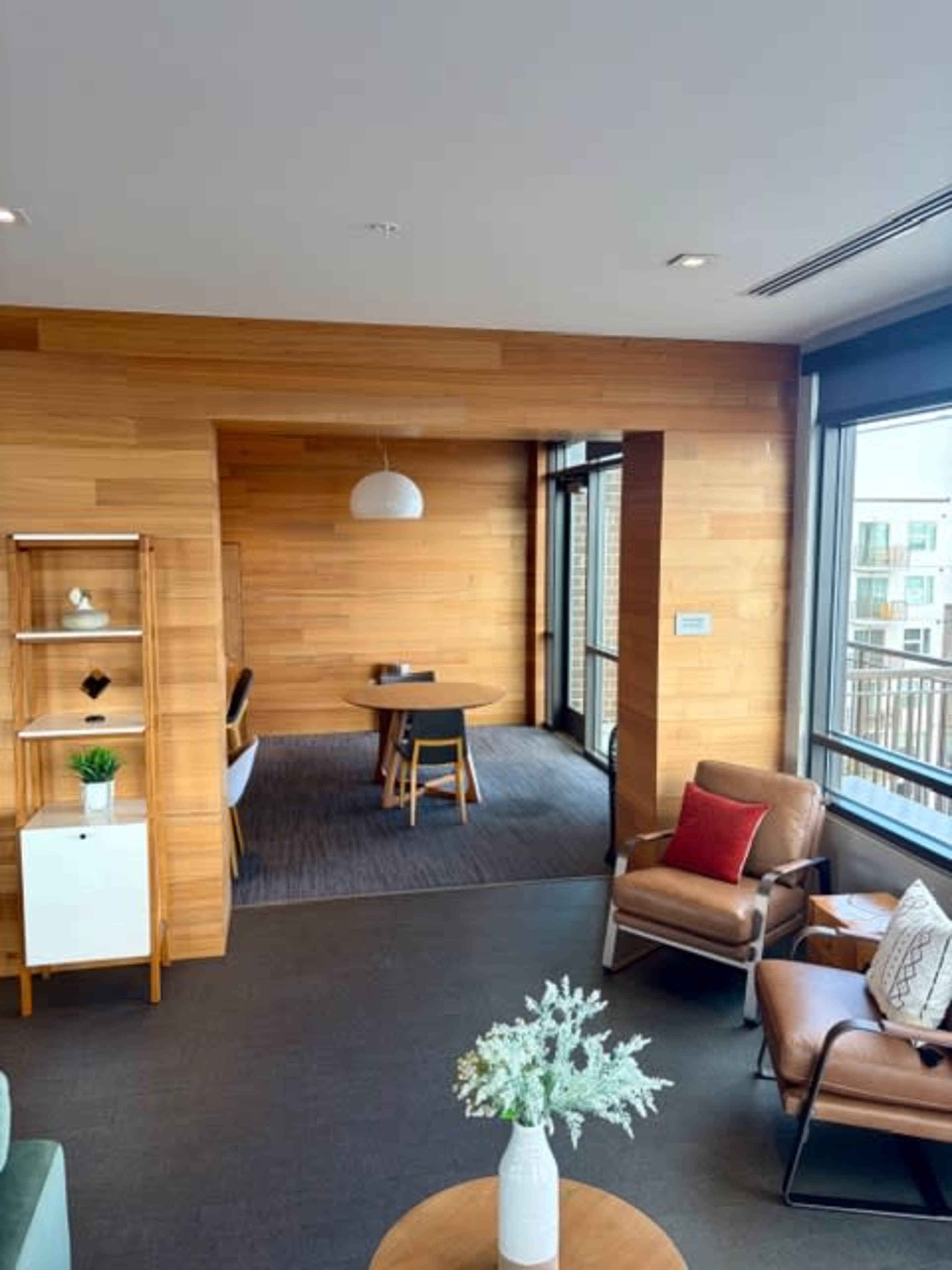 The image shows a modern interior space with wooden walls, a small dining table, and seating areas featuring leather chairs and a coffee table.