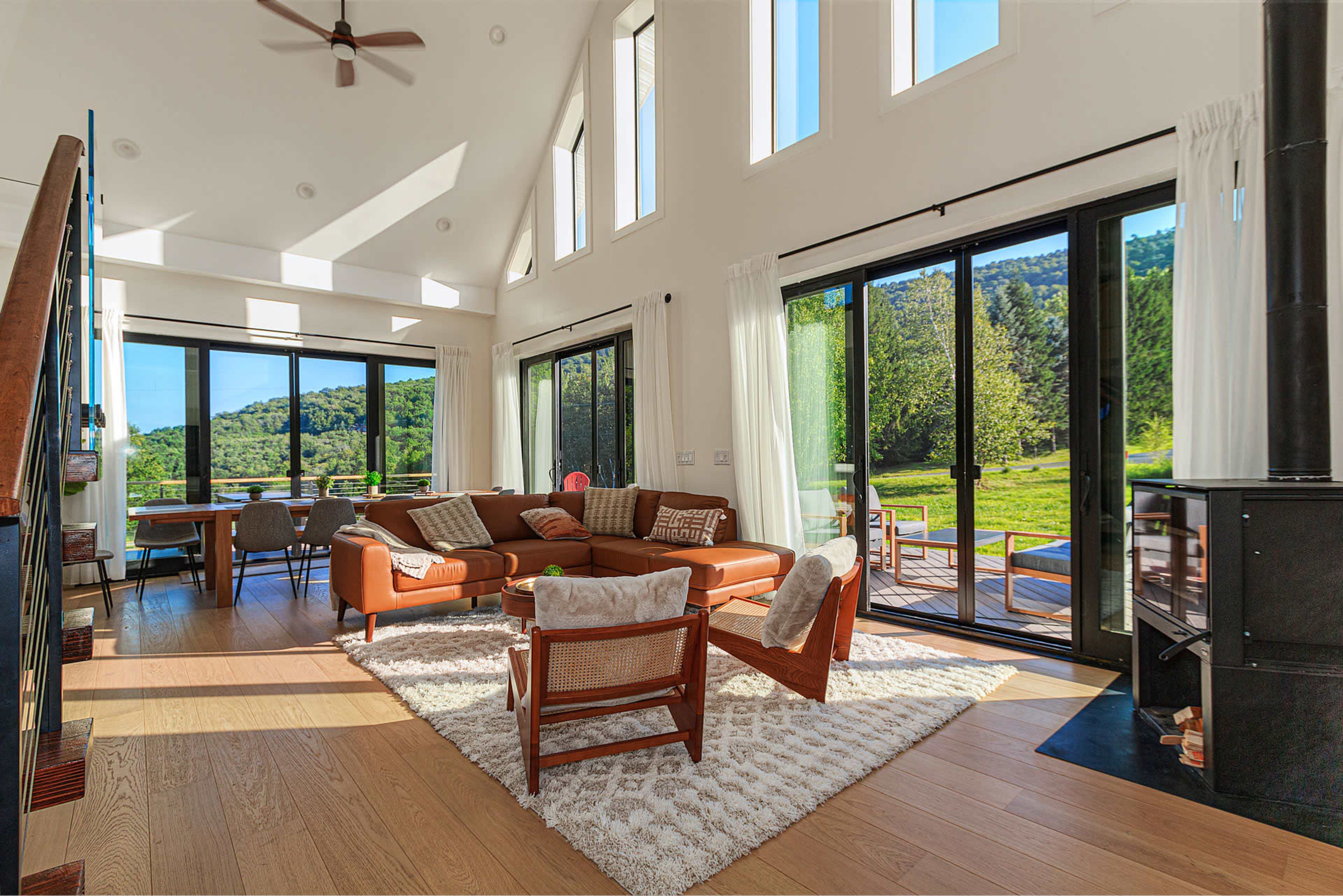 The interior of a modern living room features large windows, a brown sofa, wooden flooring, and a view of lush greenery outside.