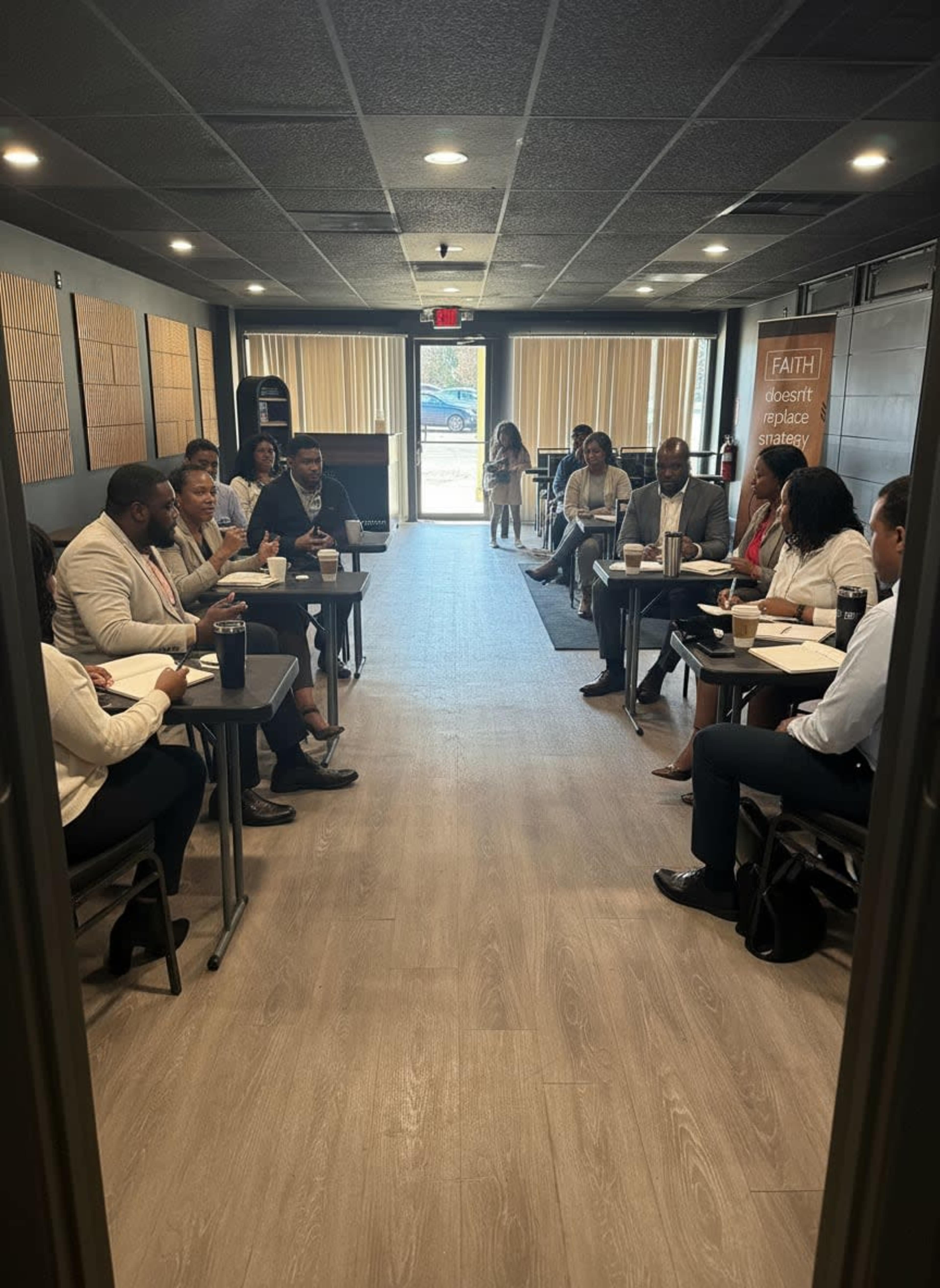 A group of people is seated around tables in a business meeting space, engaged in discussion and taking notes.