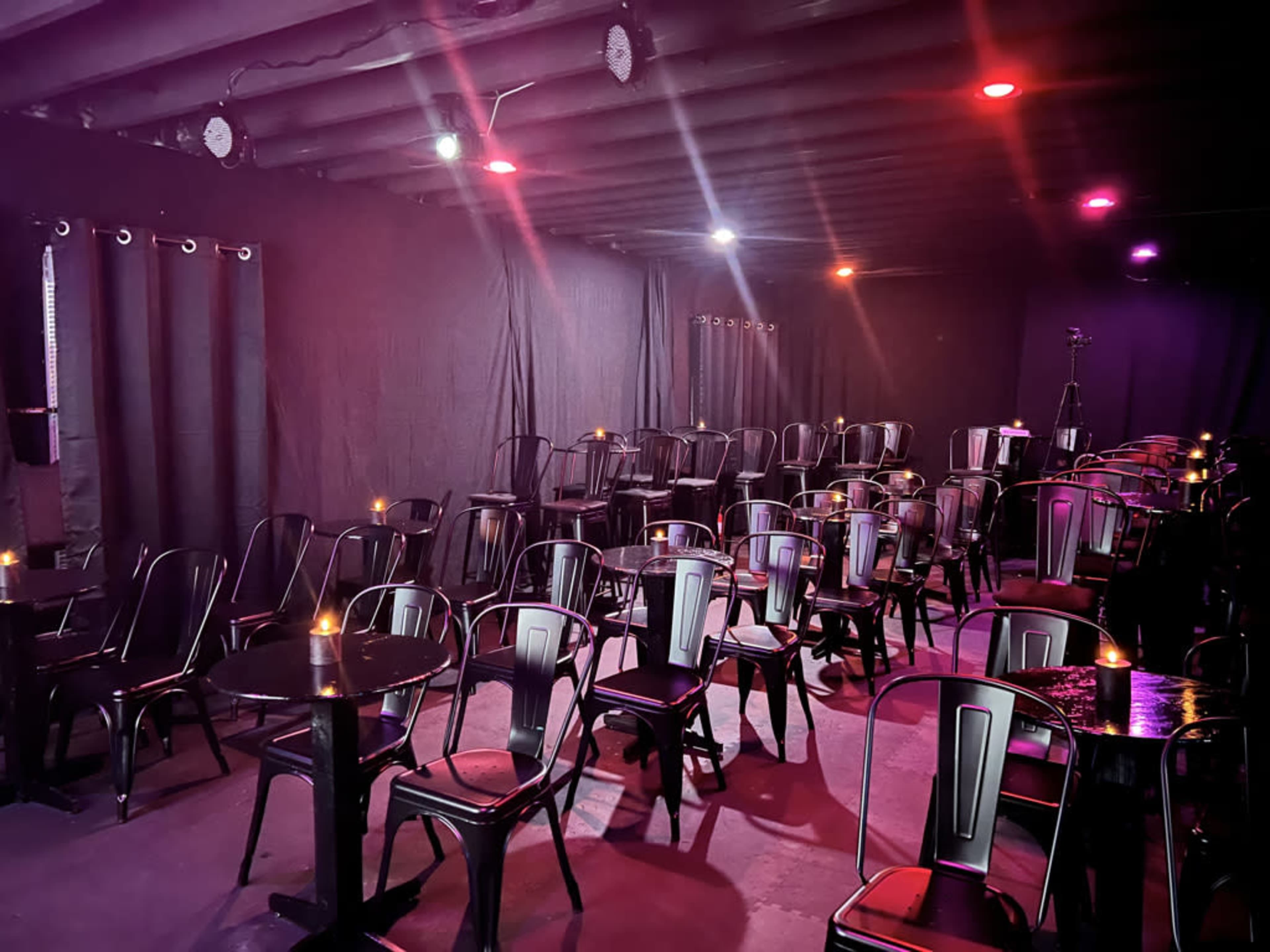 The image shows an empty performance venue with black chairs arranged around small tables, illuminated by colored stage lights.