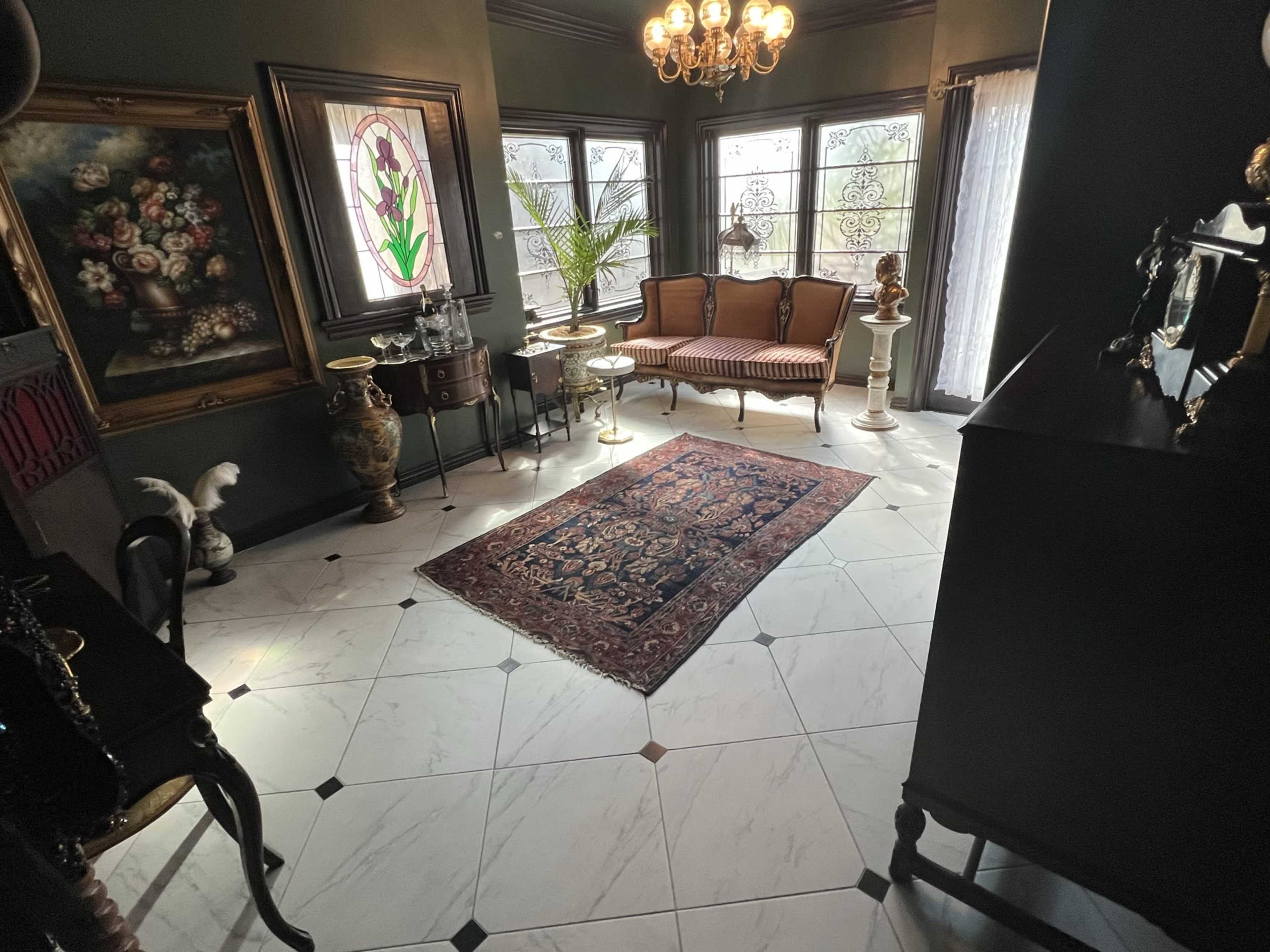 The image shows an elegantly furnished room with a patterned rug, vintage furniture, and decorative art on the walls, illuminated by natural light.