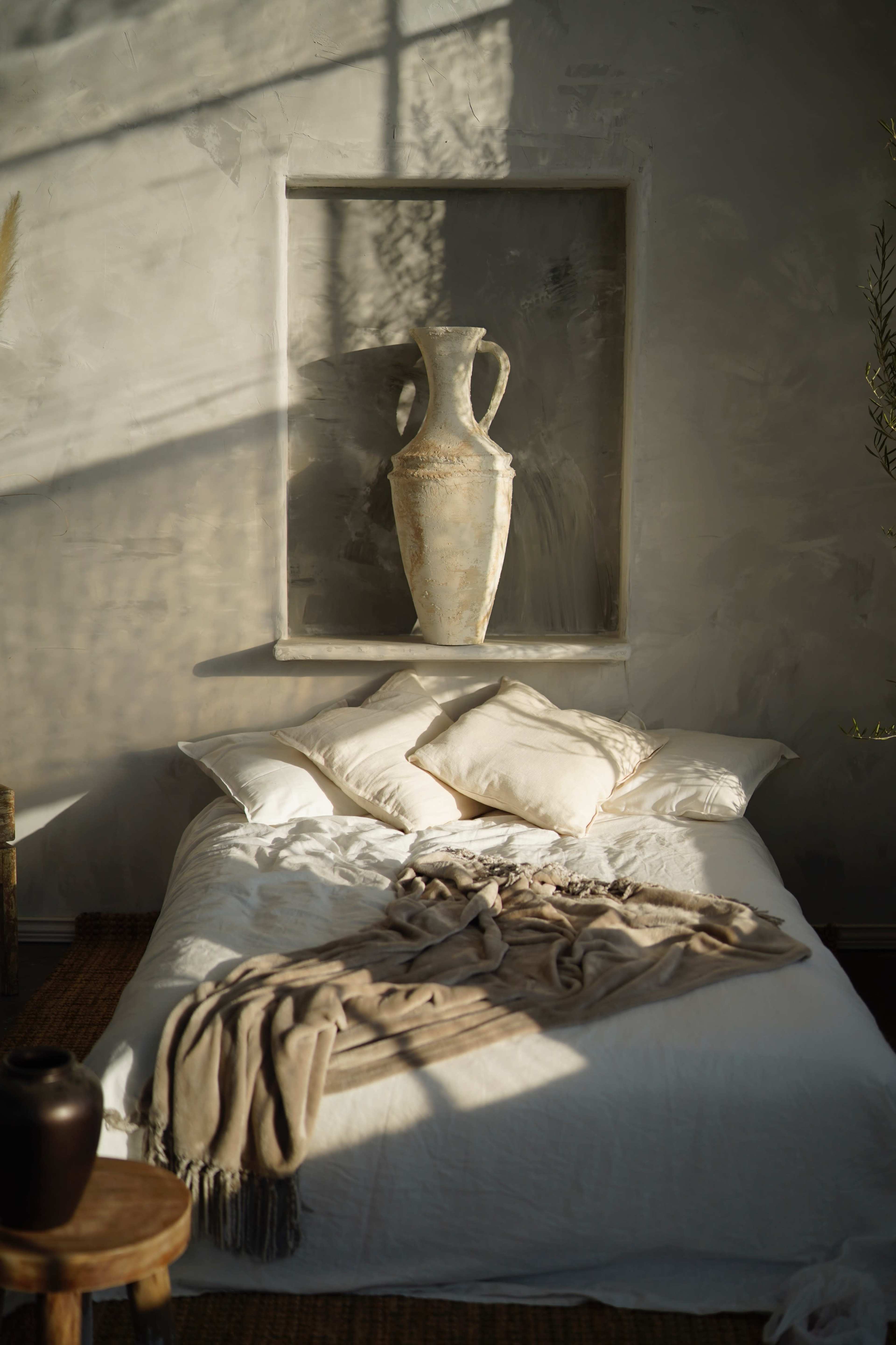 A simple bedroom features a bed with several pillows and a draped blanket, alongside a decorative ceramic vase displayed in a wall recess.