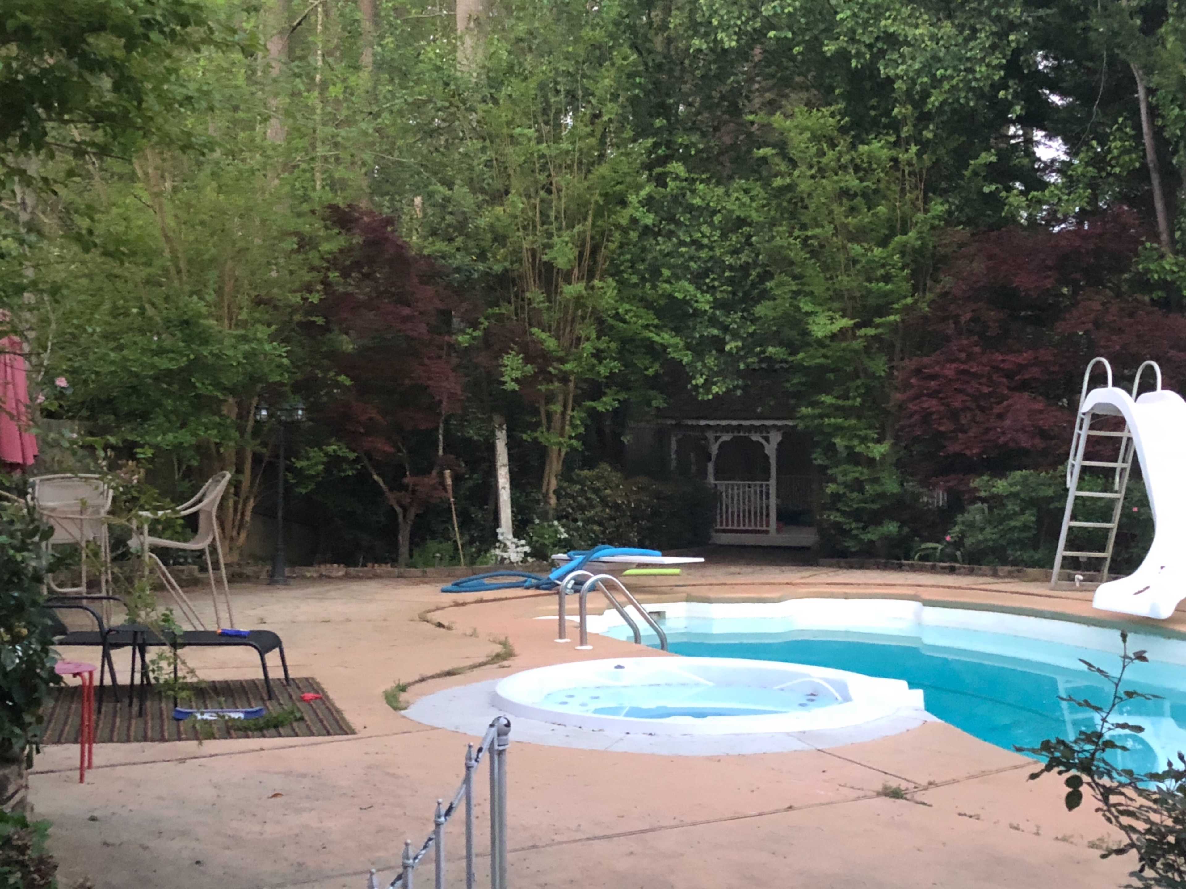A swimming pool with a slide and a hot tub is surrounded by greenery and features a gazebo in the background.