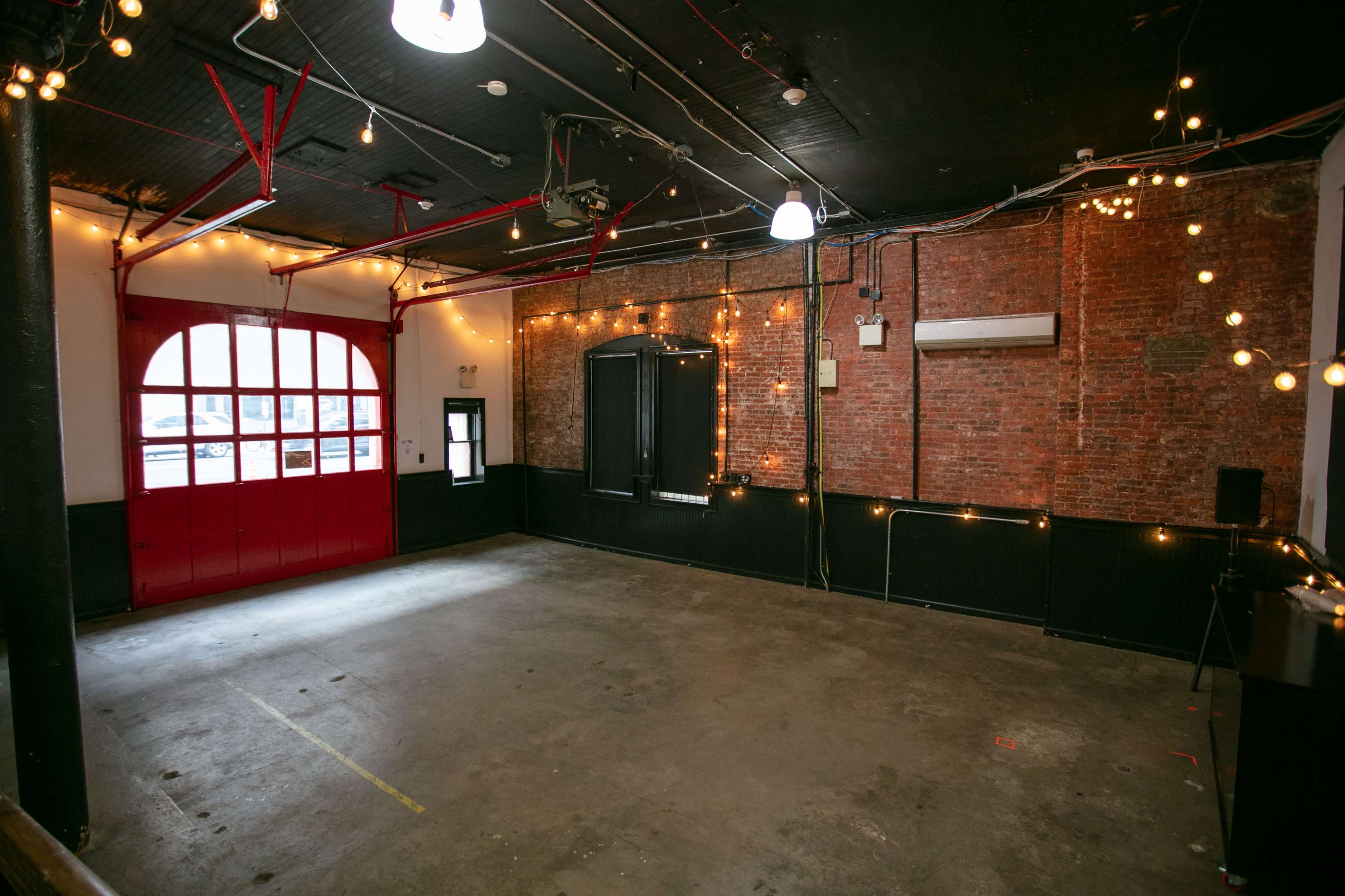 The image depicts a spacious, empty event venue featuring exposed brick walls, string lights, and large windows with a red door at one end.