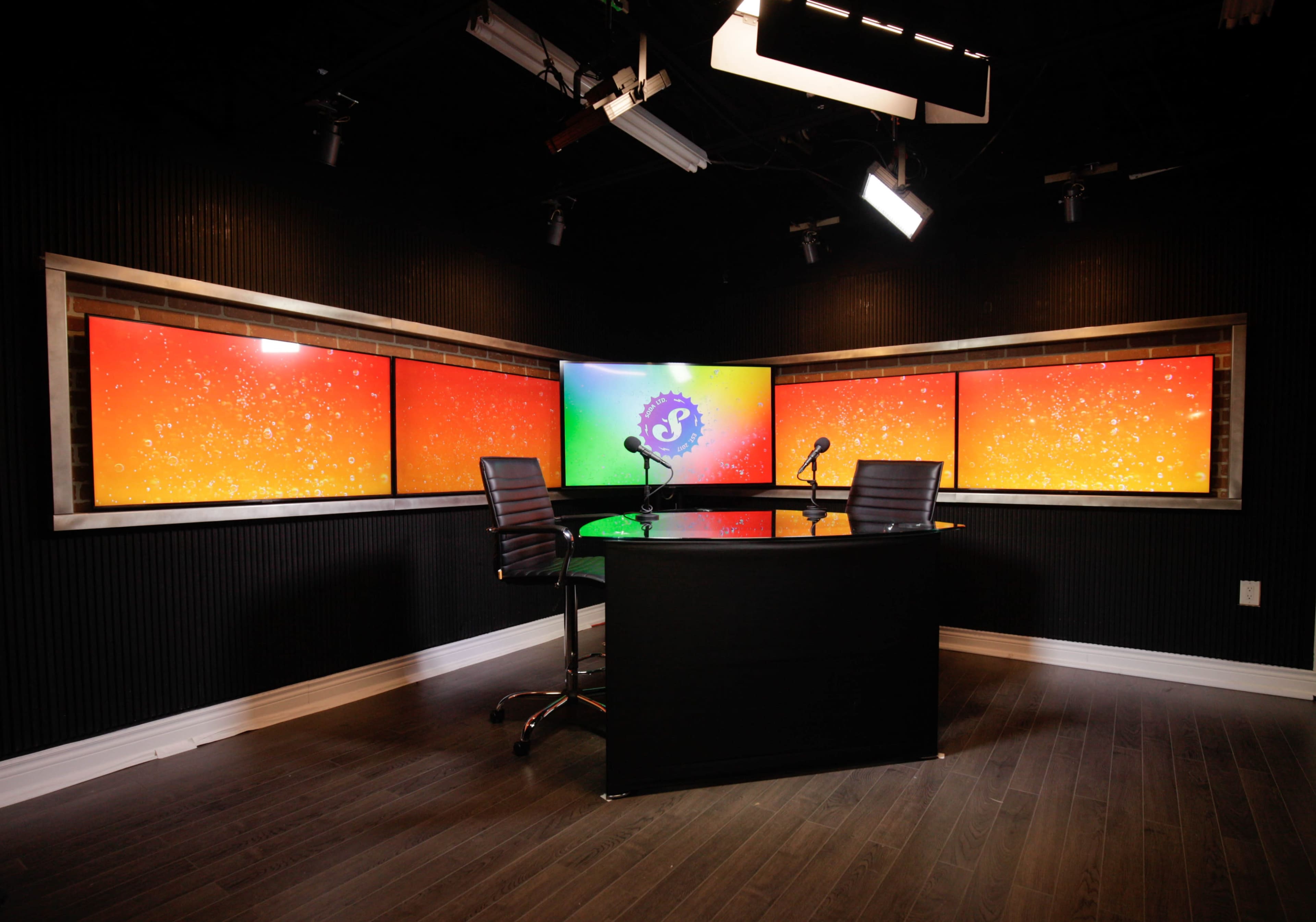 A modern studio setting features a central circular table with two chairs and multiple brightly colored screens in the background.