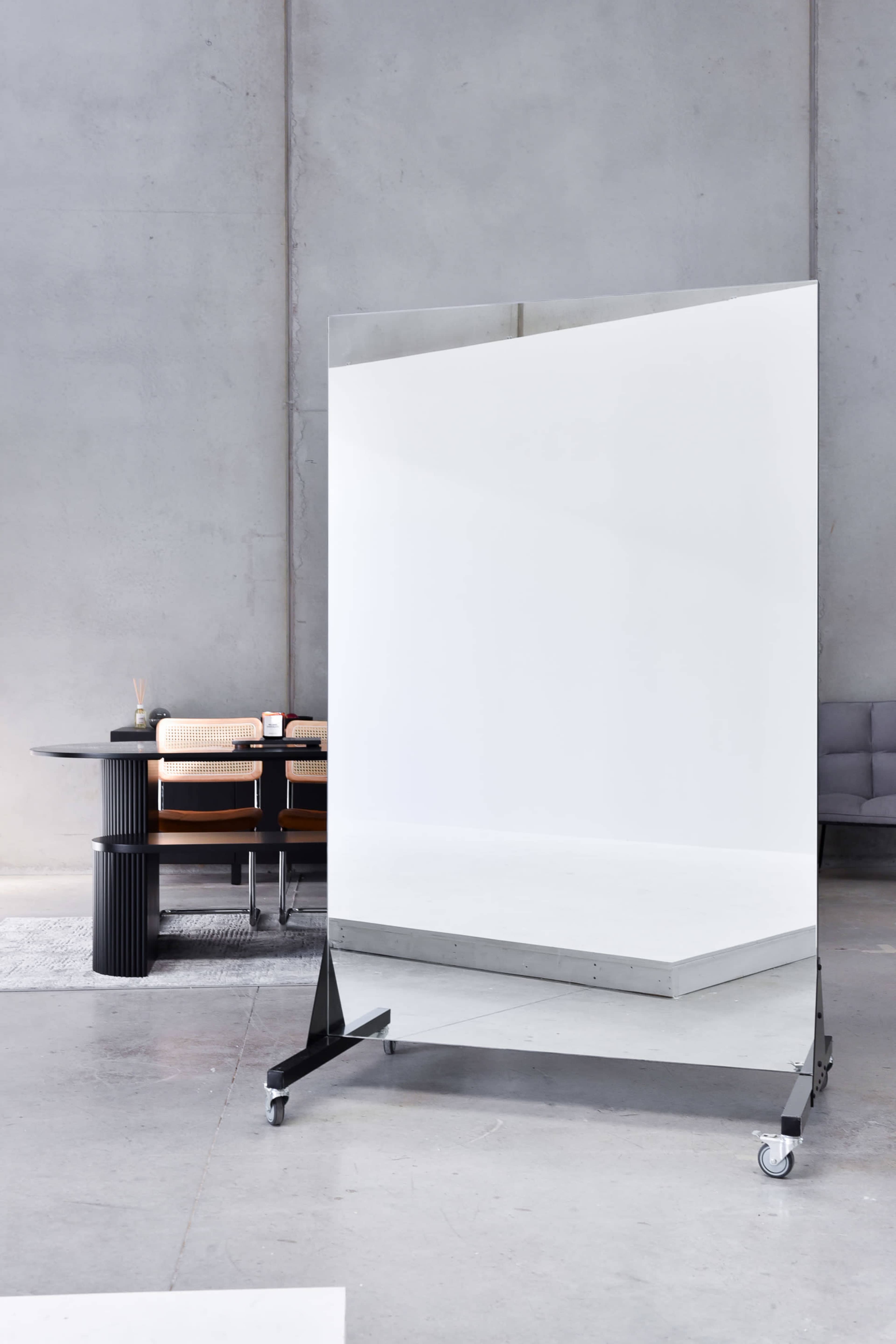 A large, freestanding mirror on wheels stands in a minimalist, industrial-style room featuring a dining table and chairs in the background.