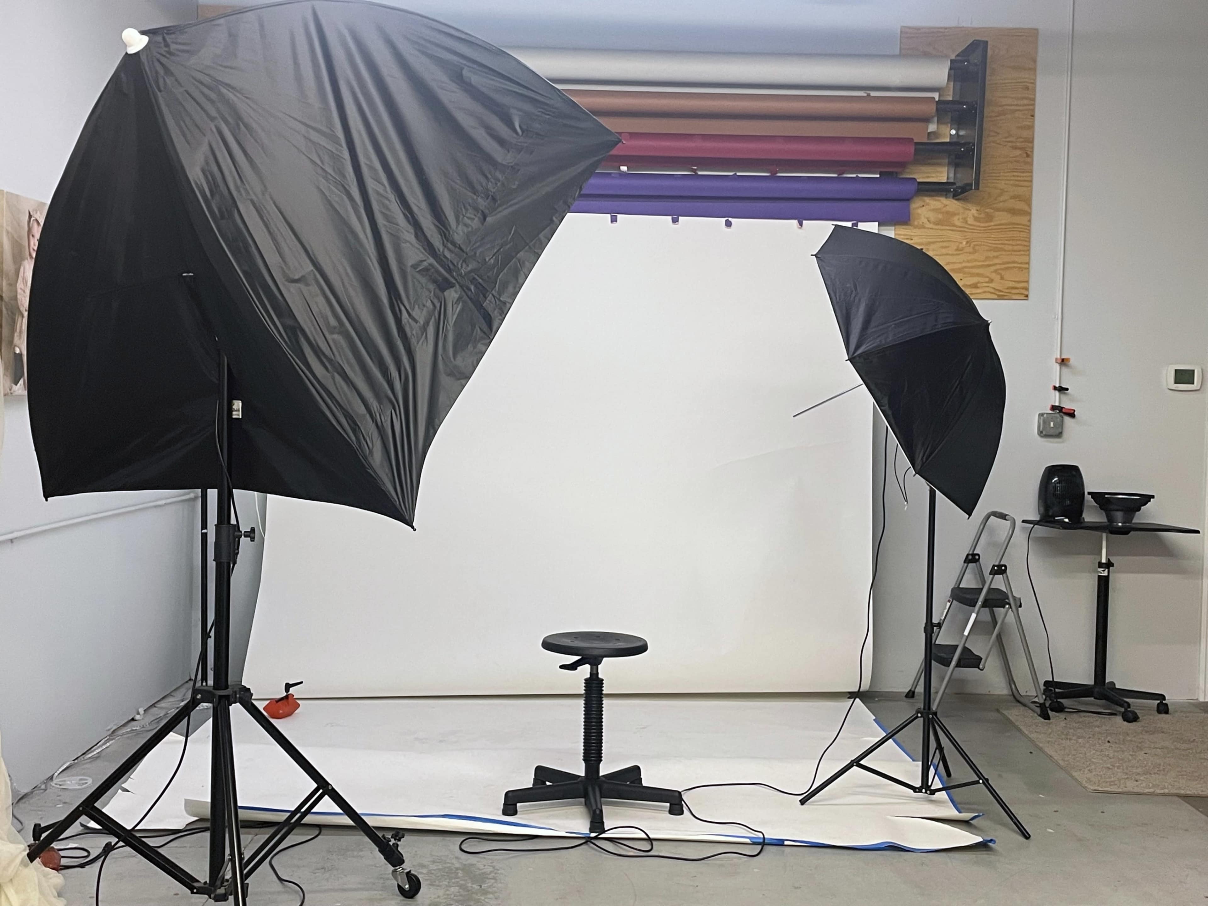 The image shows a photography studio setup with two umbrellas, a stool in the center, and various backgrounds hung on the wall.