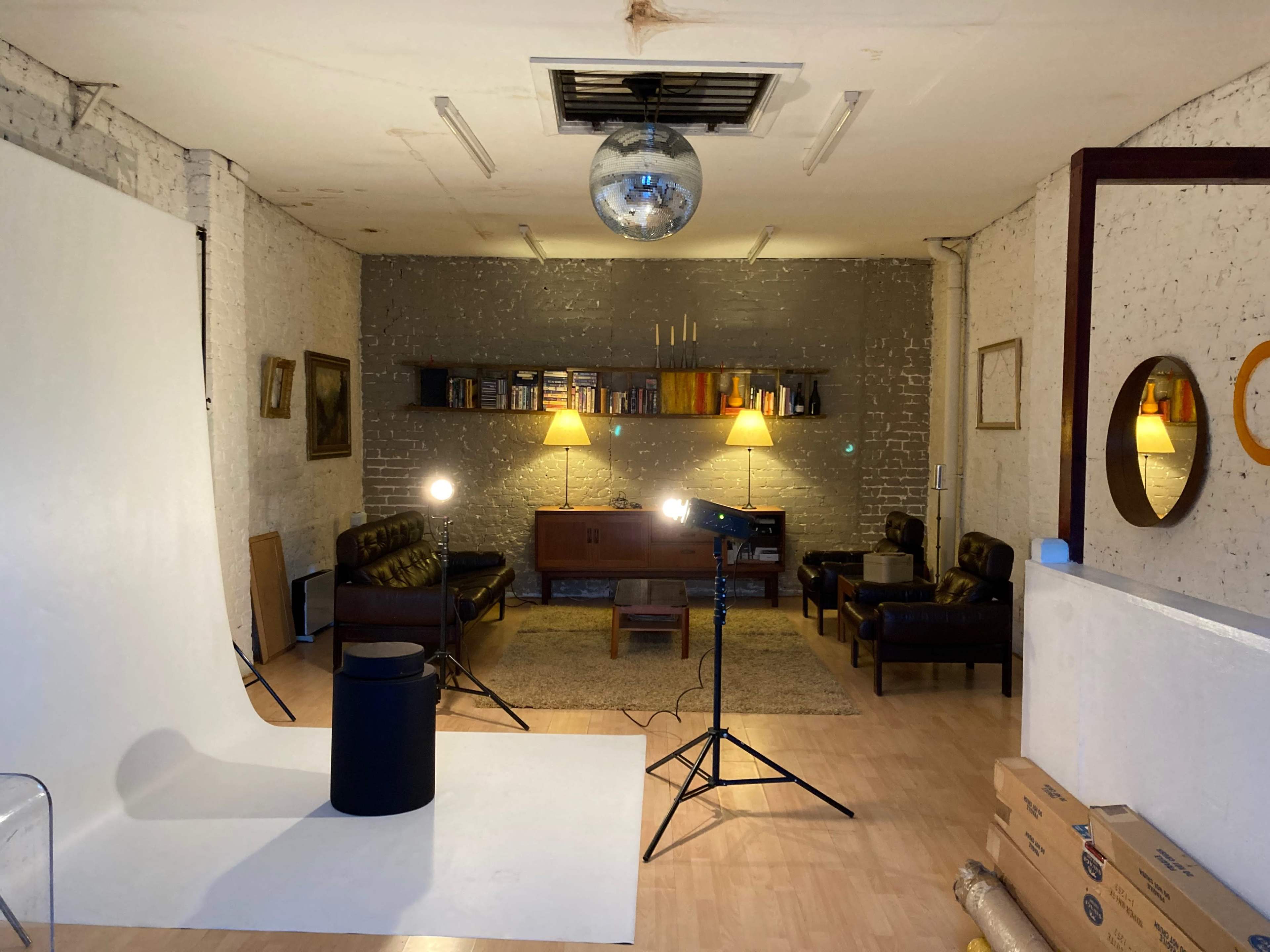 The image depicts a minimalist studio space with a white backdrop, black furniture, two standing lights, and a disco ball hanging from the ceiling.