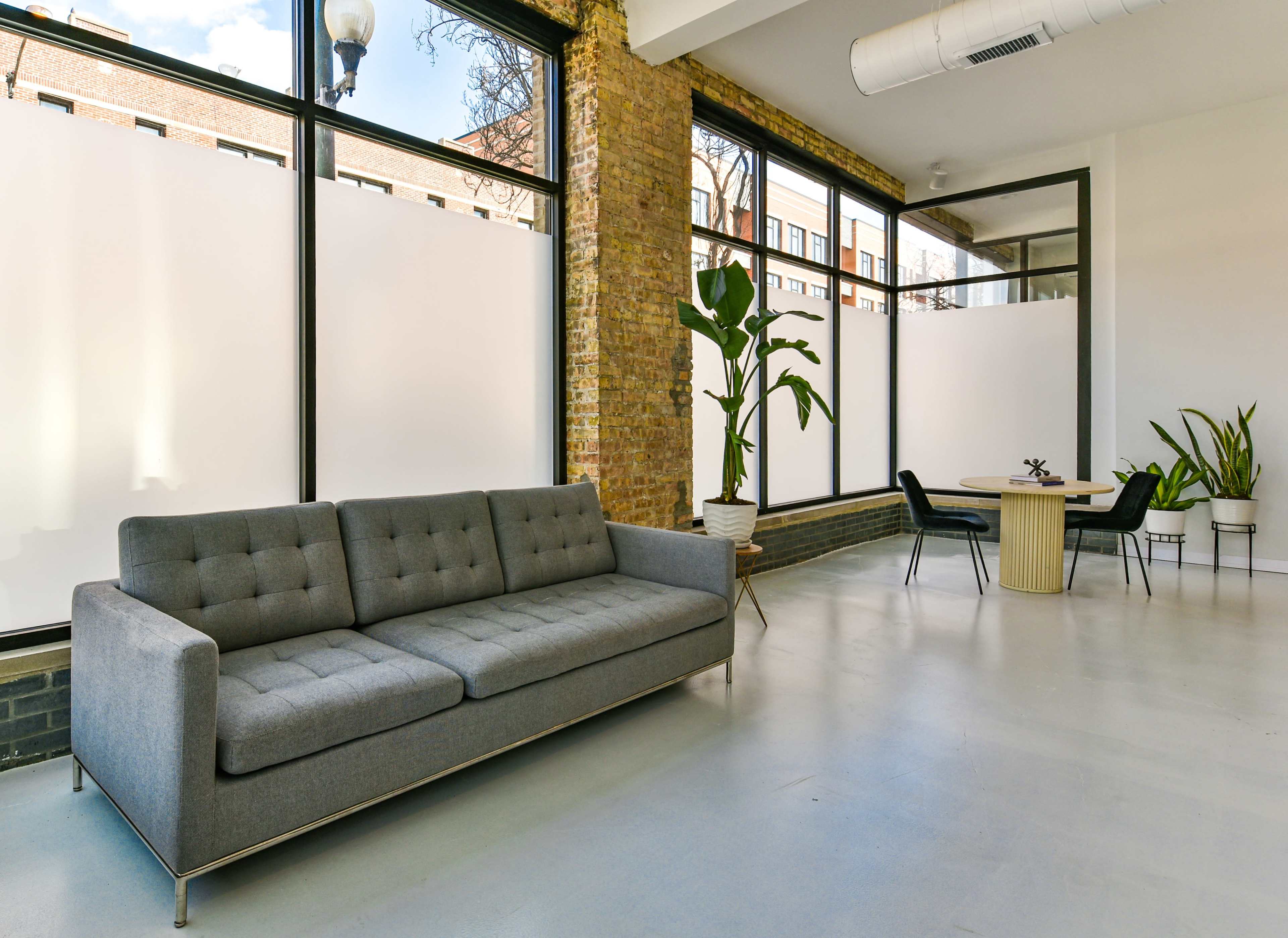 A spacious room with large windows, featuring a gray sofa and a round wooden table surrounded by chairs, alongside potted plants.