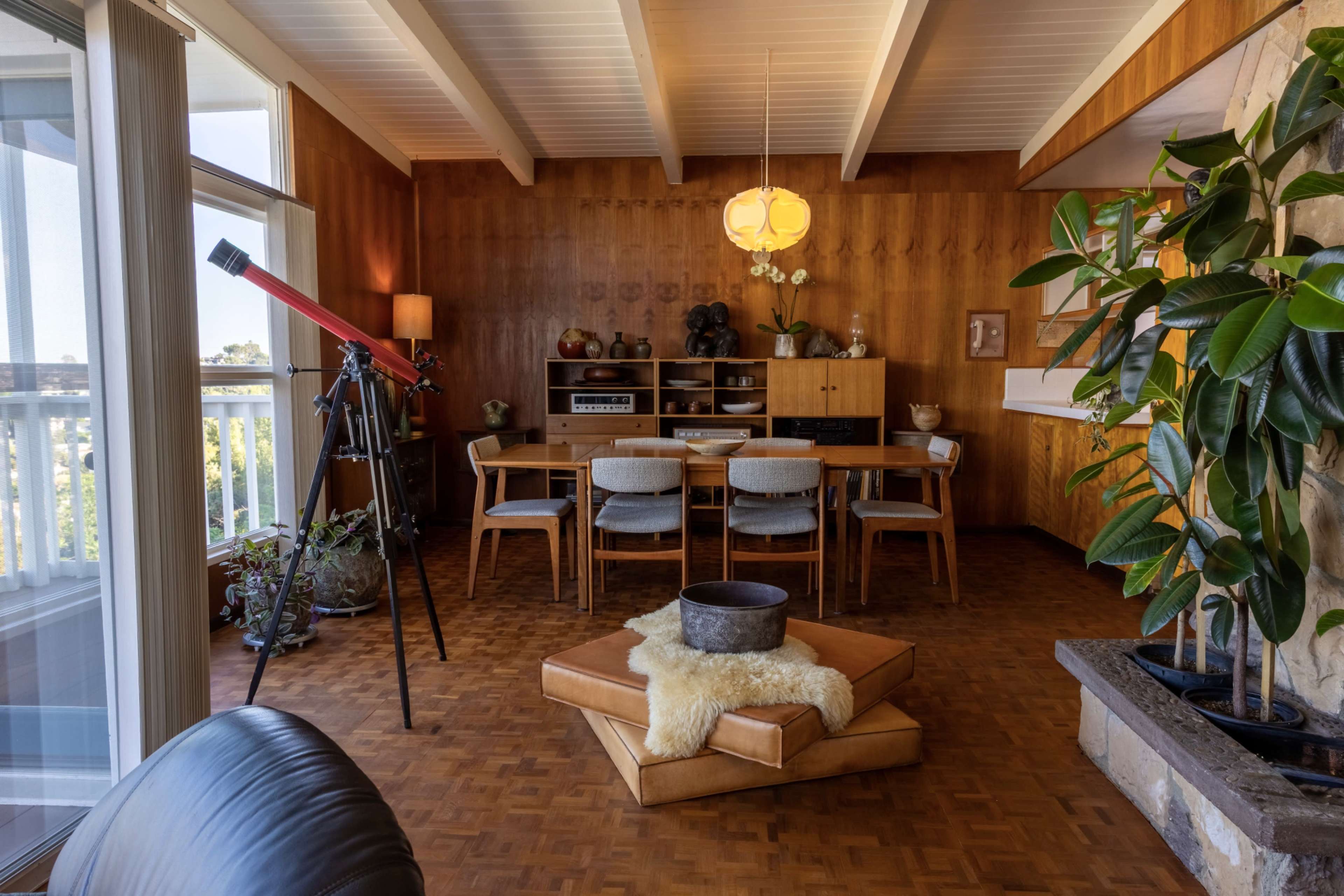 The image shows a mid-century modern dining area with a wooden table and chairs, a telescope, and large windows that provide ample natural light.