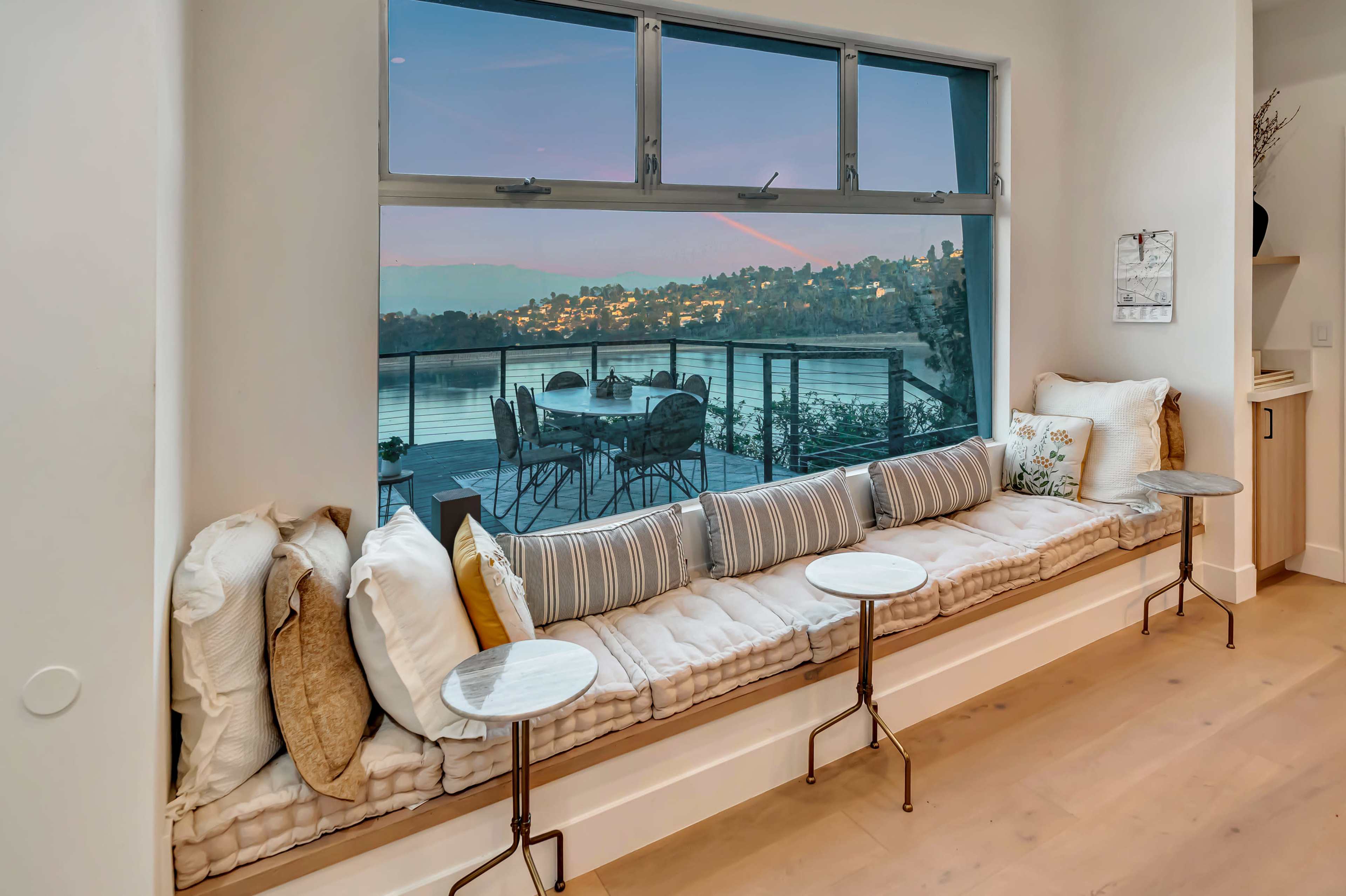 The image shows a cozy window seat with cushions and small tables, overlooking a scenic view of a lake and city lights in the distance.