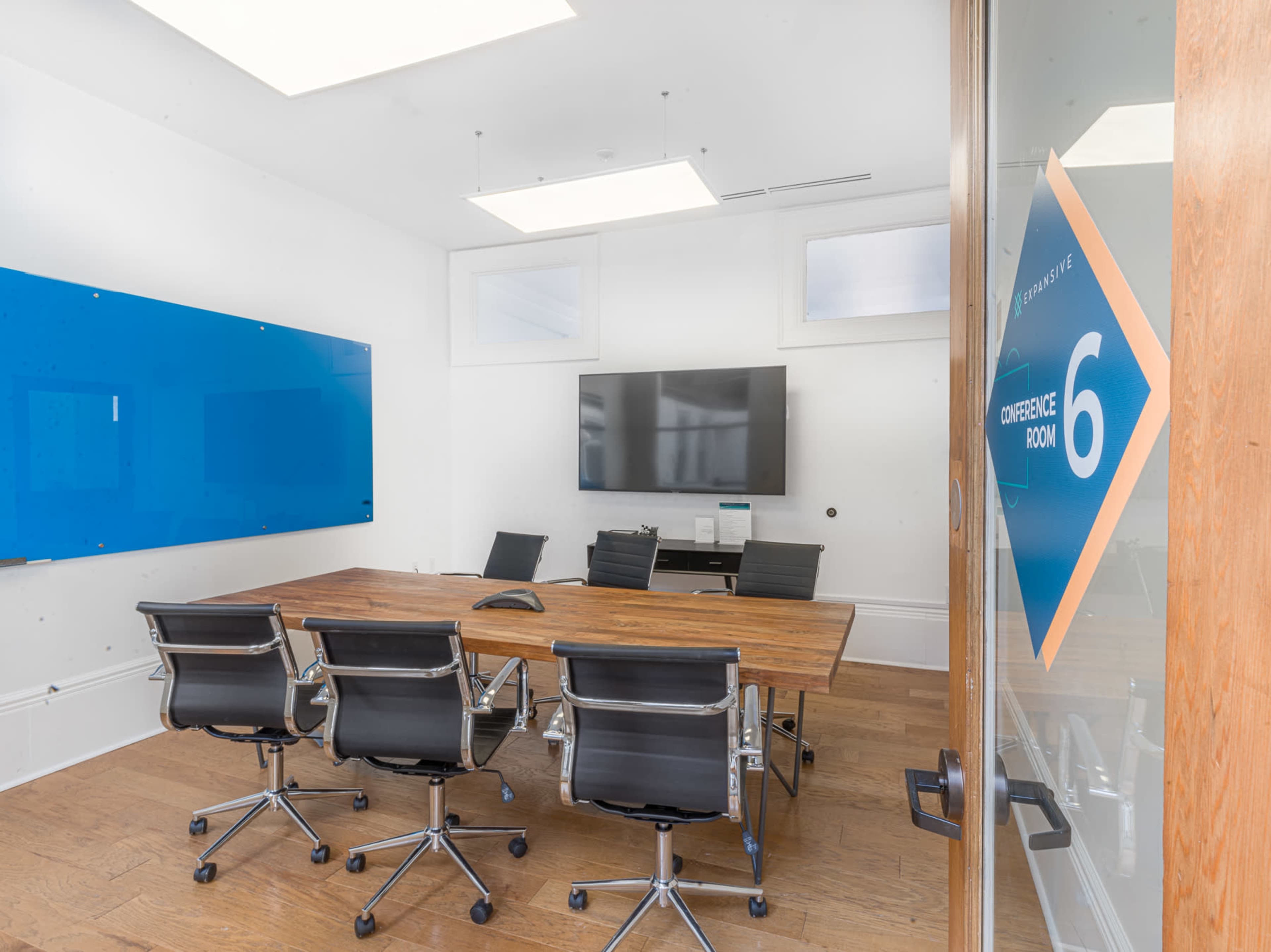 The image shows a conference room with a large wooden table, several black swivel chairs, a television on the wall, and a blue whiteboard.