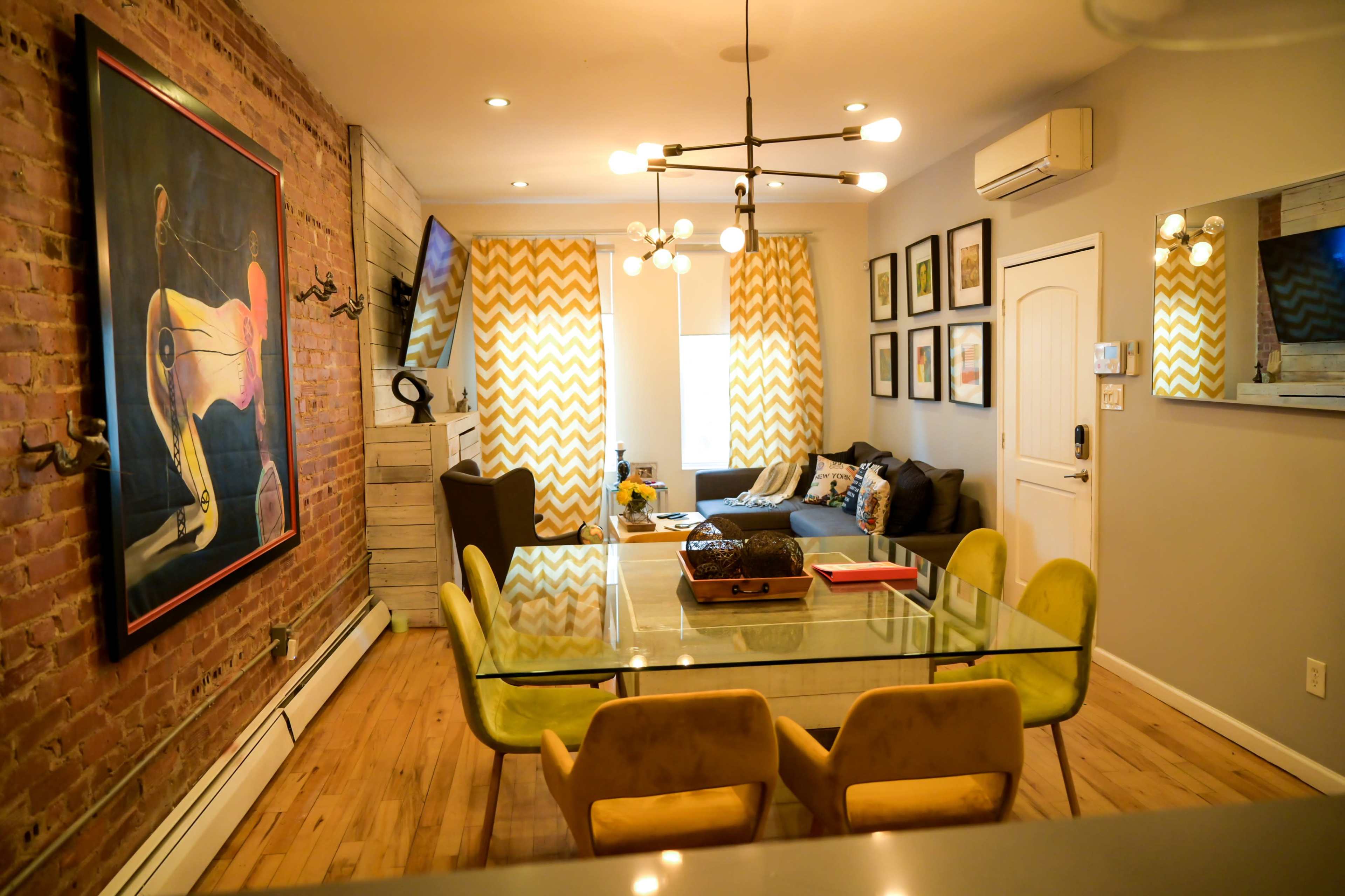 A modern living and dining area featuring a glass table surrounded by yellow chairs, a sectional sofa, and decorative wall art against a brick backdrop.