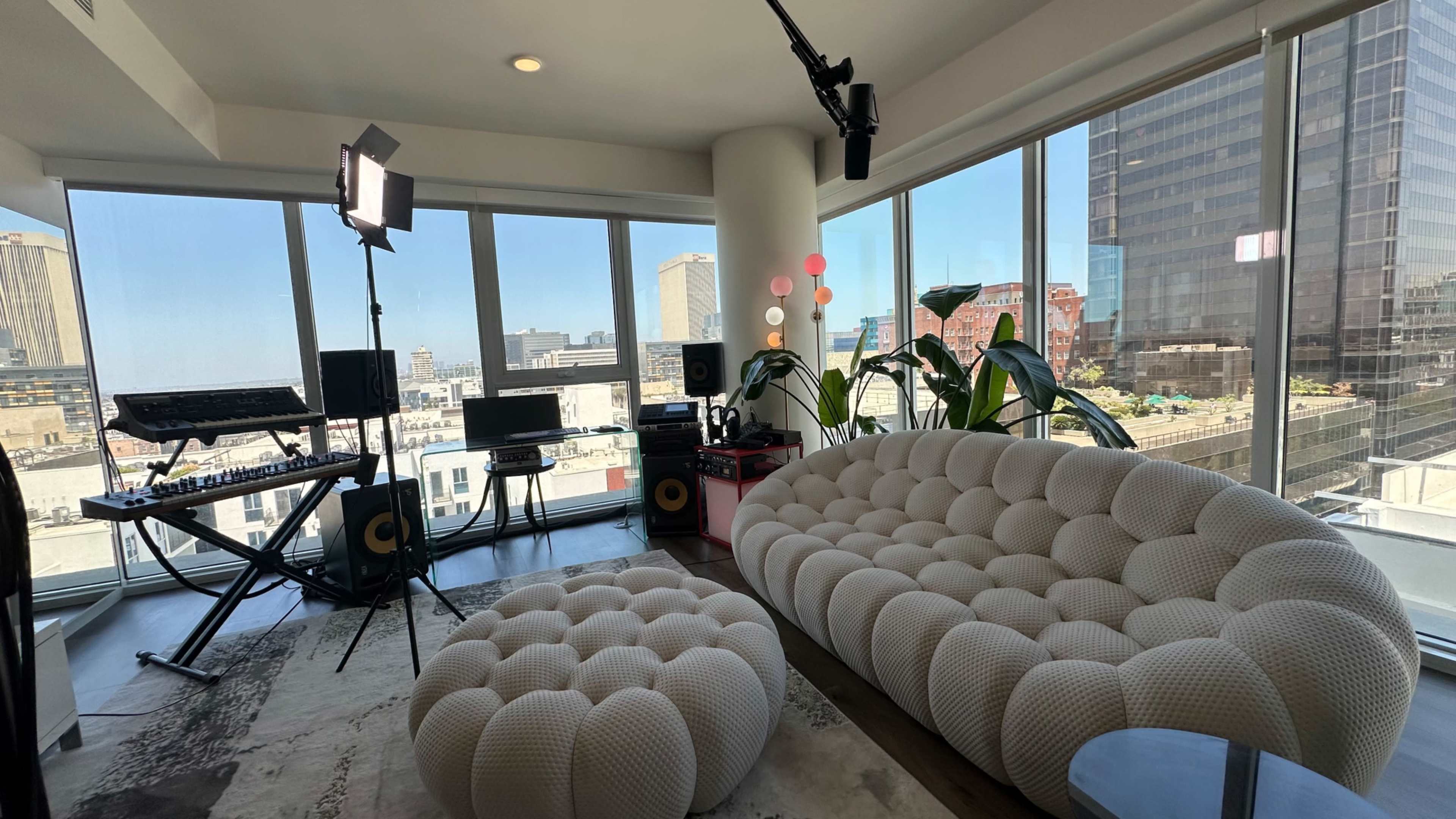 A modern interior features a white, tufted sofa and ottoman, with a music production setup near large windows offering a city view.