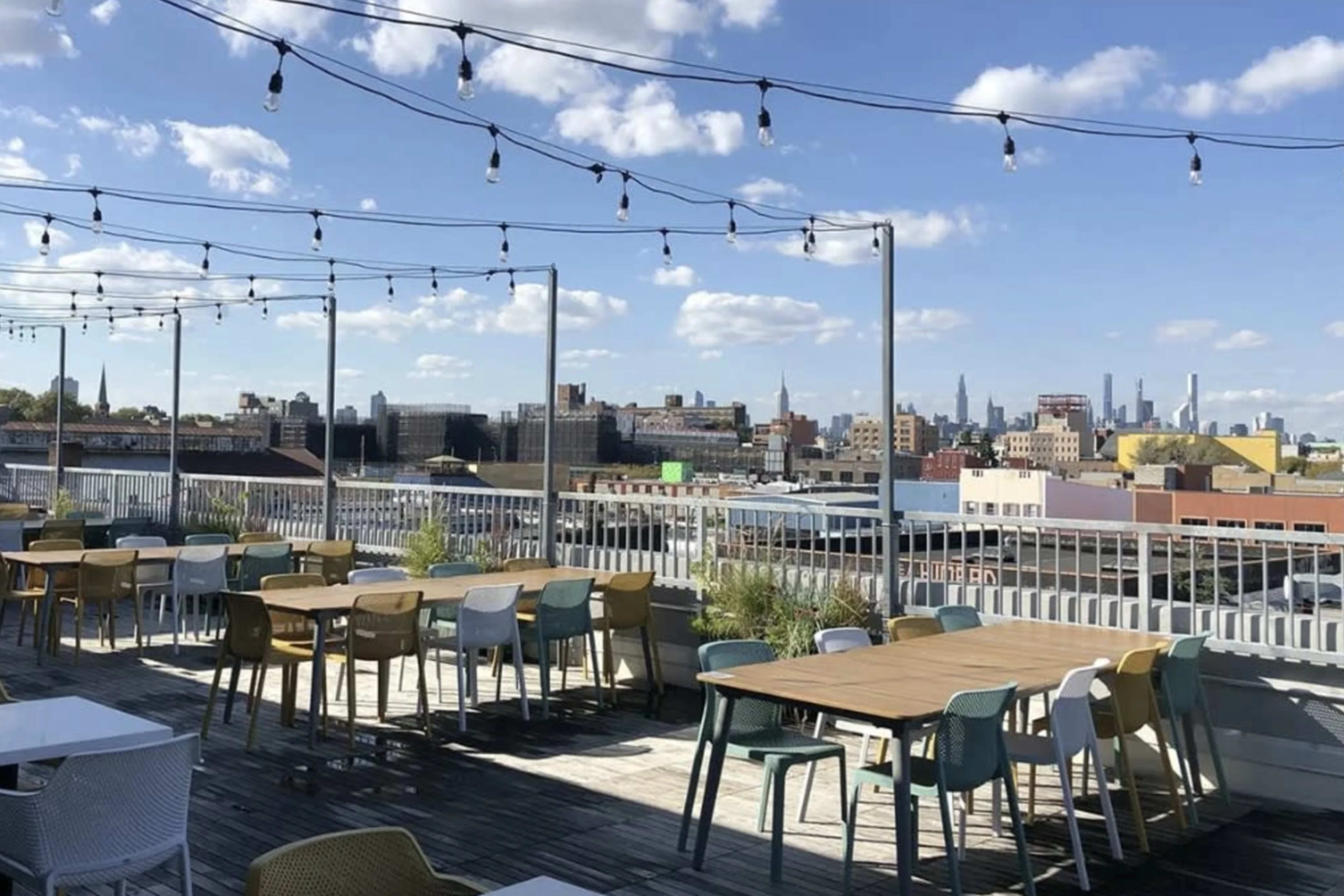 A rooftop terrace features several tables and chairs with a view of the city skyline under a clear blue sky.