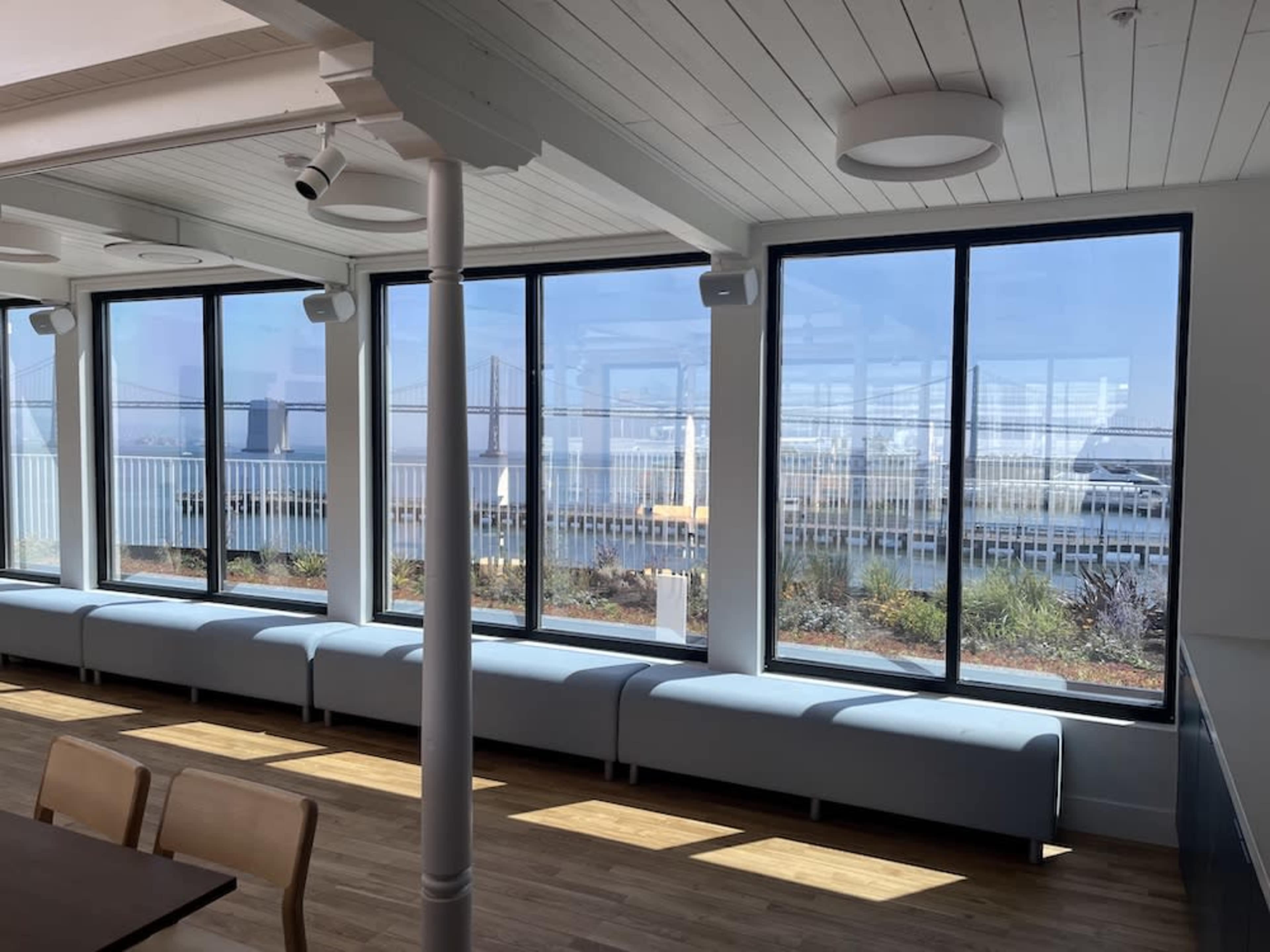 A bright interior space with large windows overlooking a body of water and a bridge in the background.