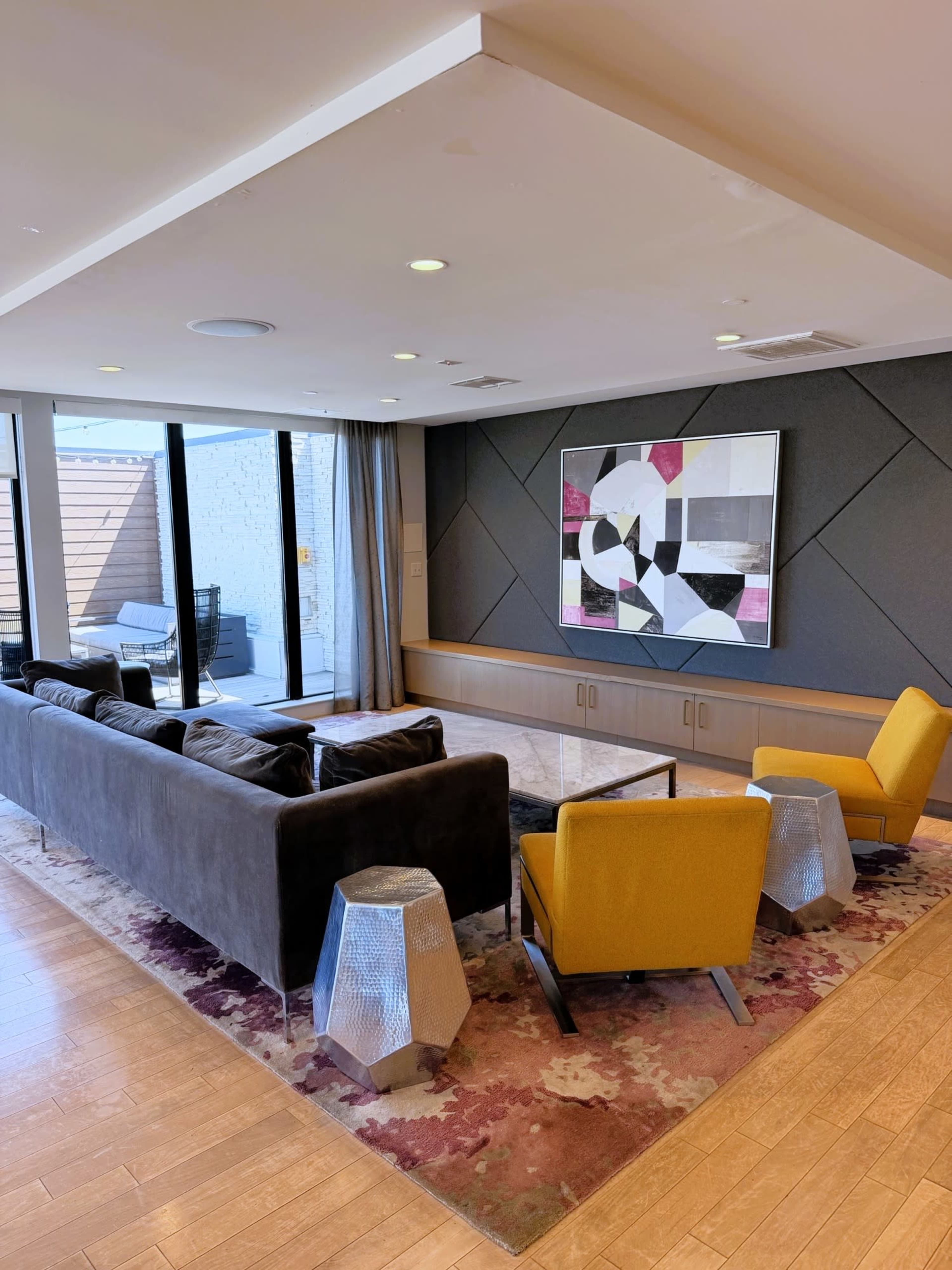 The image shows a modern lounge area featuring a gray sofa, two yellow armchairs, a glass coffee table, and a large abstract artwork on the wall, with natural light coming in through large windows.