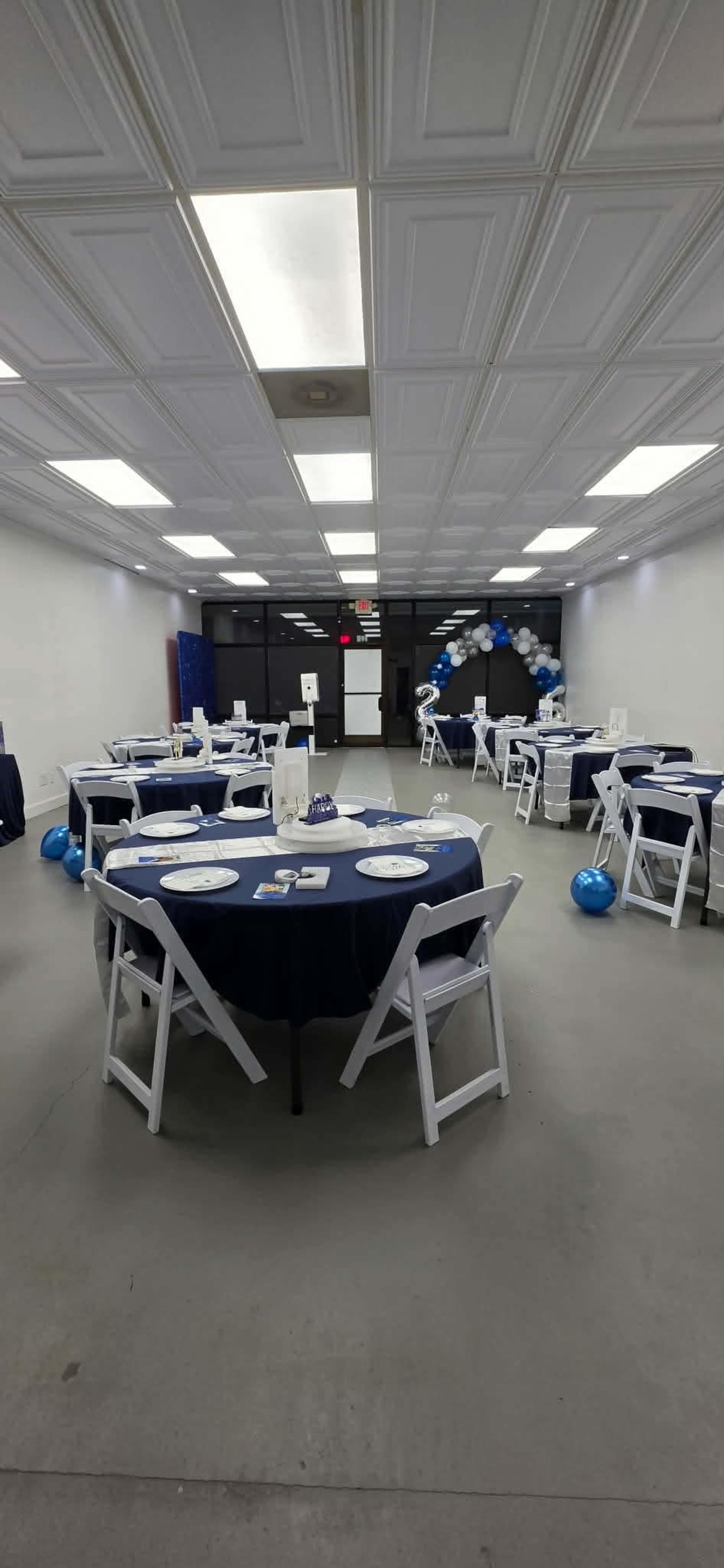 The image shows a spacious event room with several round tables covered in blue tablecloths, set with plates and decorations, and a balloon arch in the background.