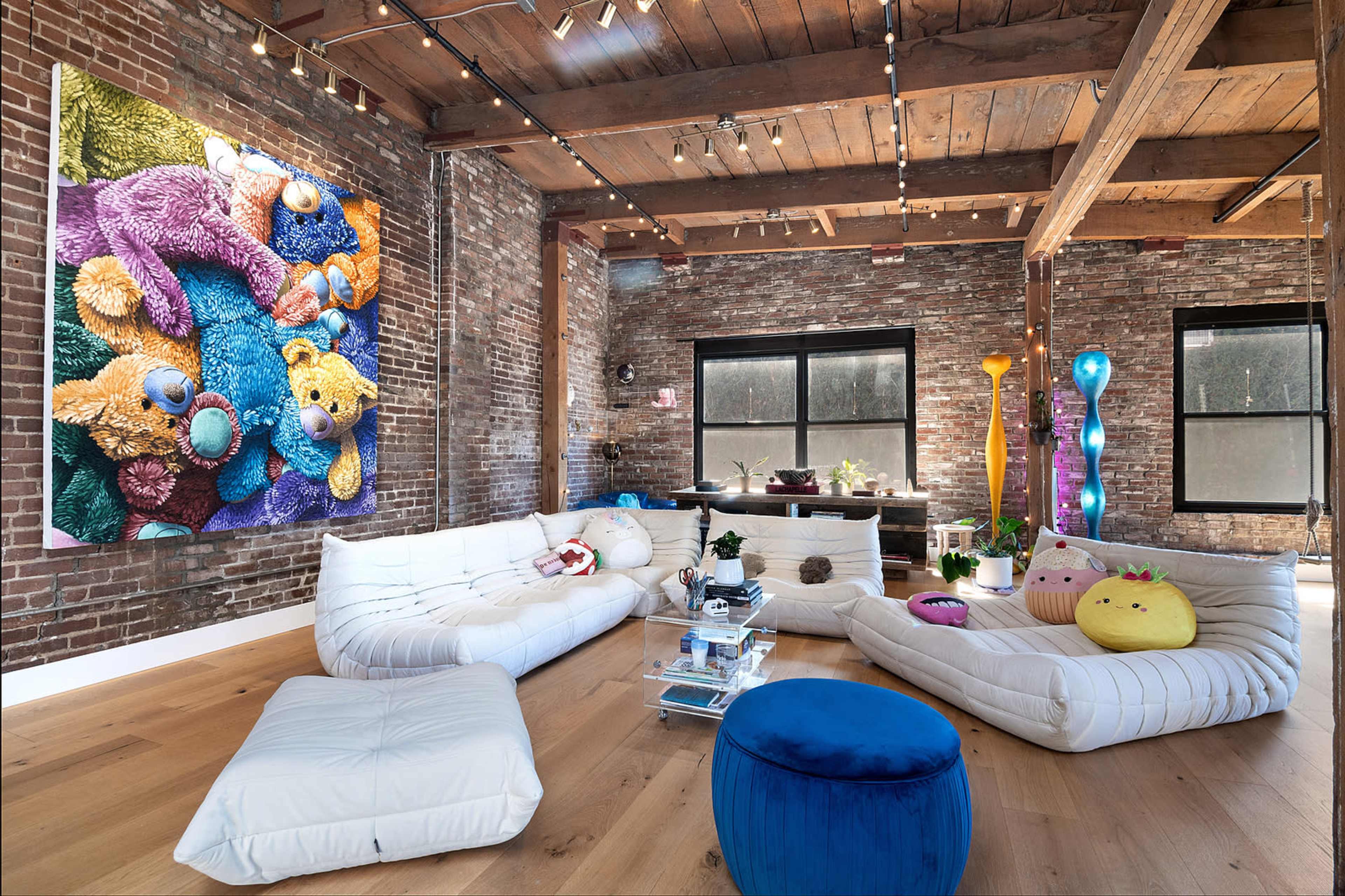 A spacious living area with exposed brick walls, a large colorful bear-themed artwork, and various seating options, including white lounge cushions and a blue ottoman.