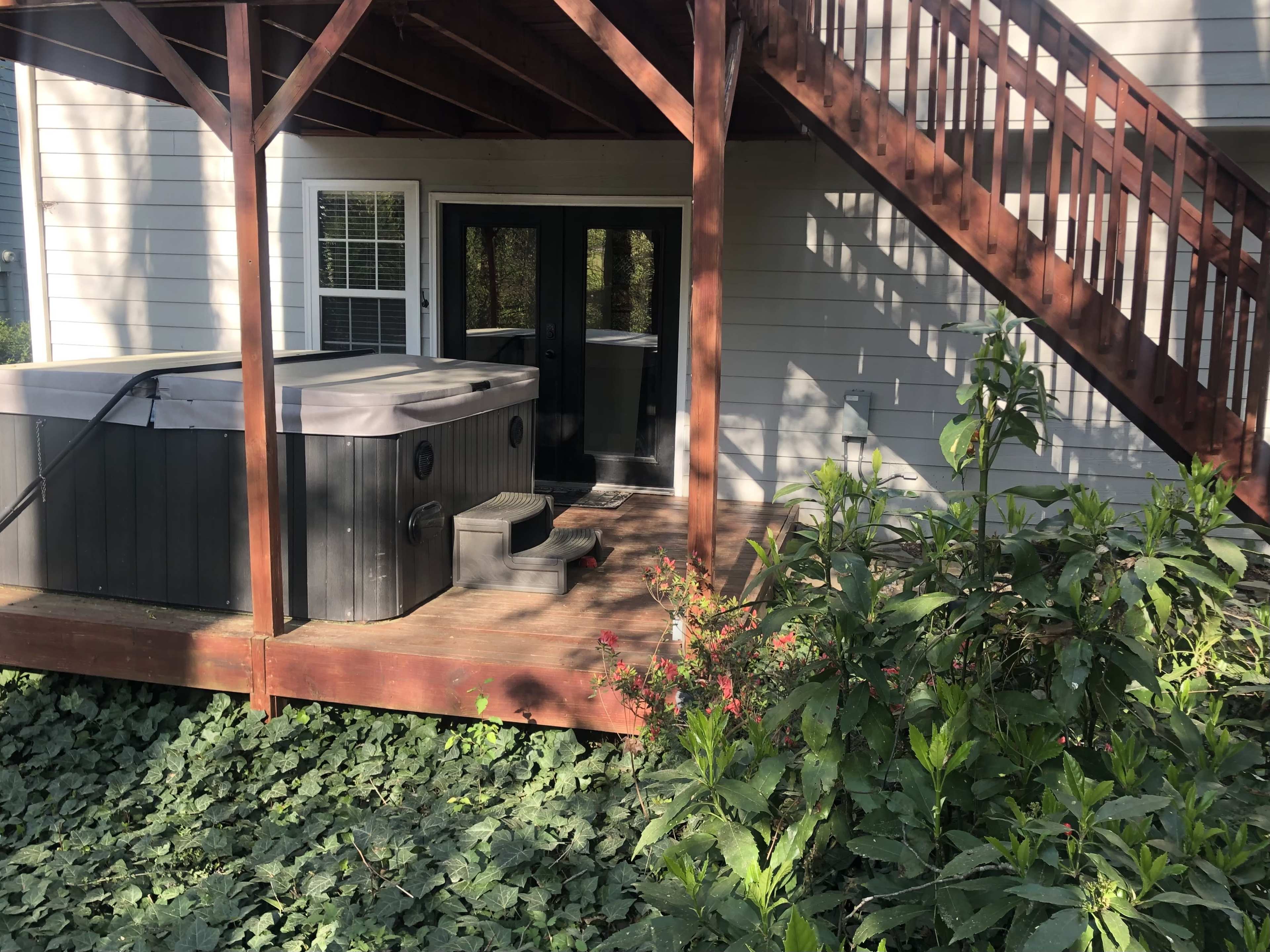 A wooden deck featuring a hot tub is located beneath a set of stairs at the back of a house.