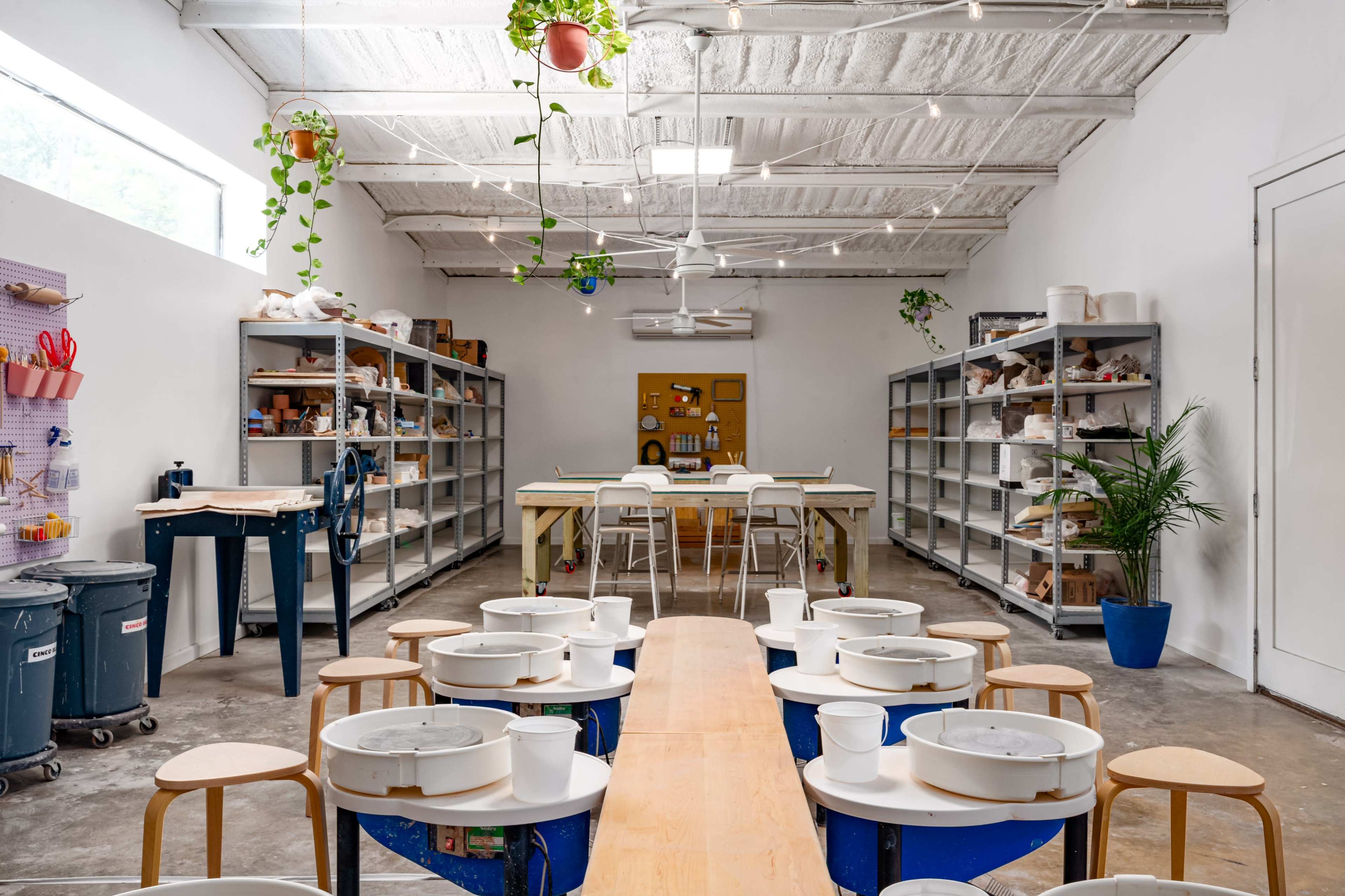 A spacious craft studio features long wooden tables with white buckets arranged in rows, surrounded by shelves and plants.