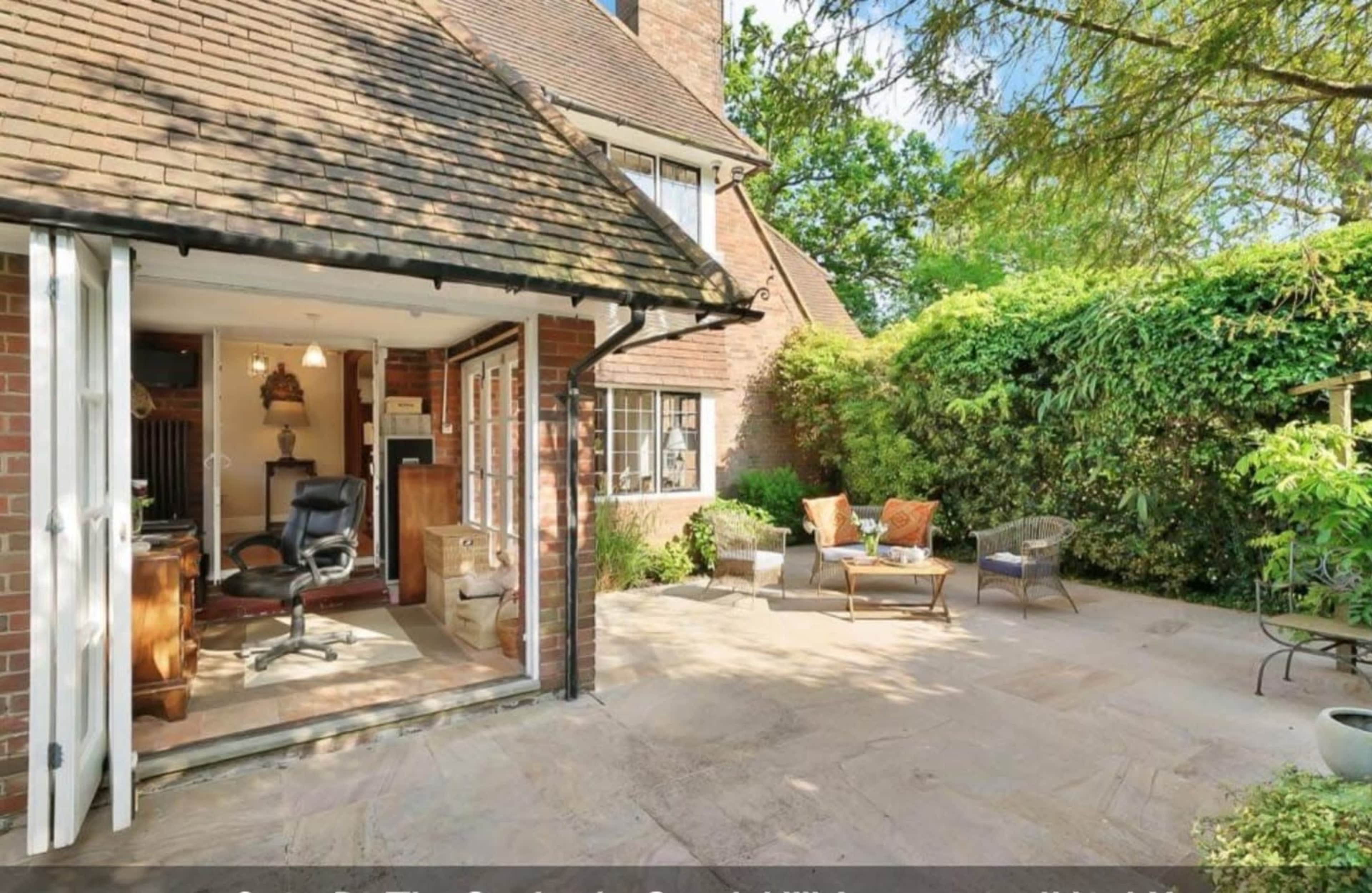 A stone patio area with outdoor seating is adjacent to a brick house surrounded by greenery.