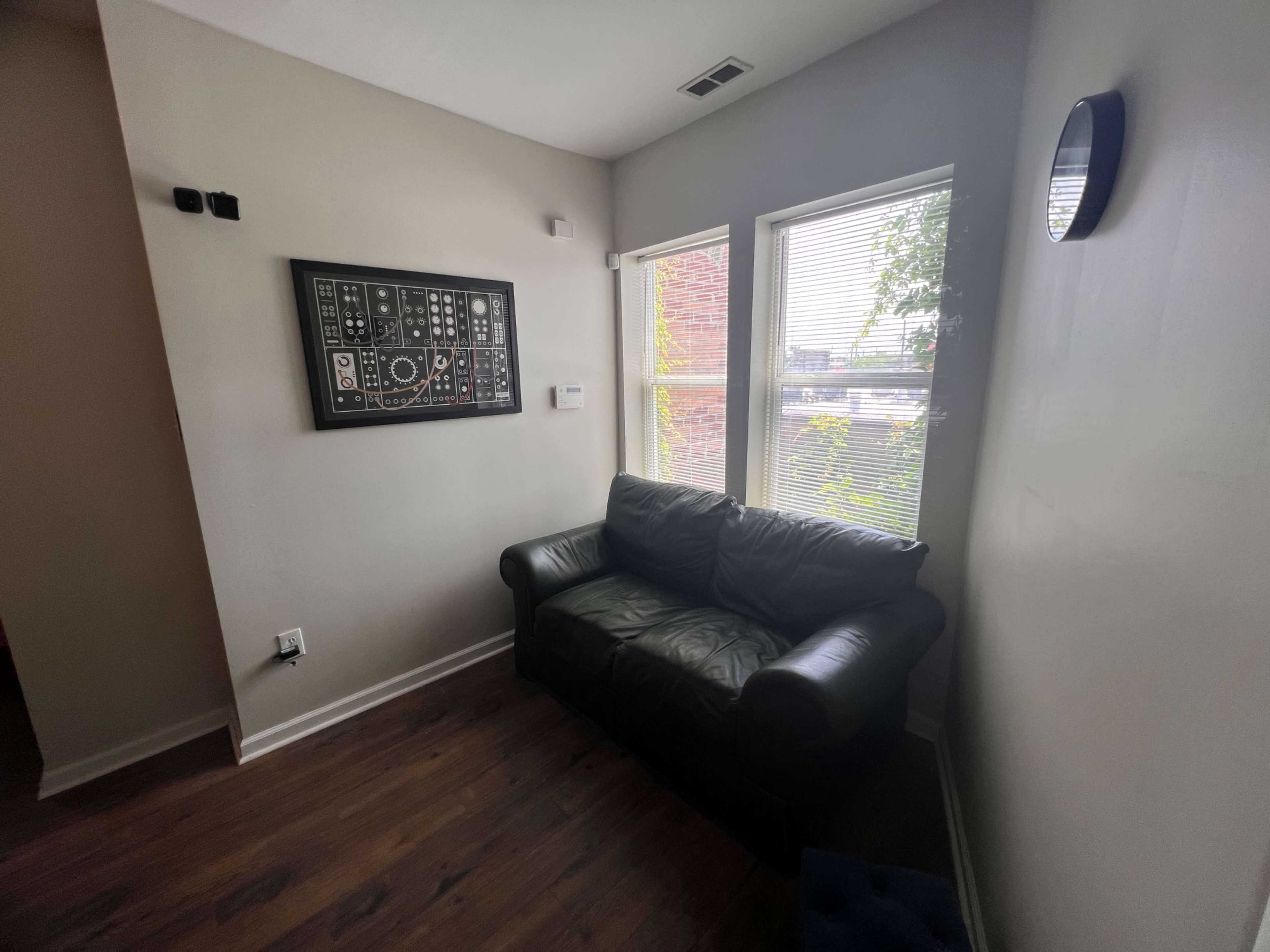 A small living area with a black leather couch, two windows, and a framed wall art piece.